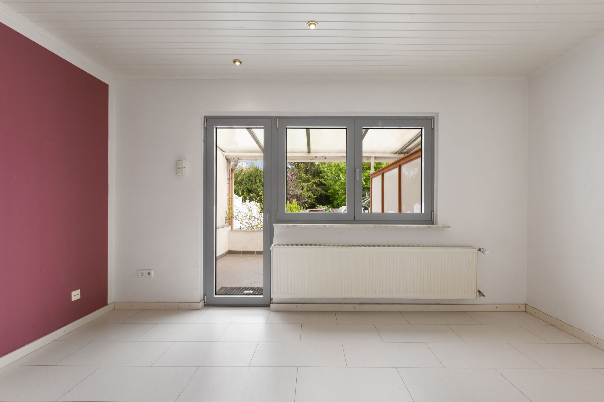 Helles Zimmer mit weißem Fliesenboden, großer Fensterfront und Blick auf eine Terrasse.