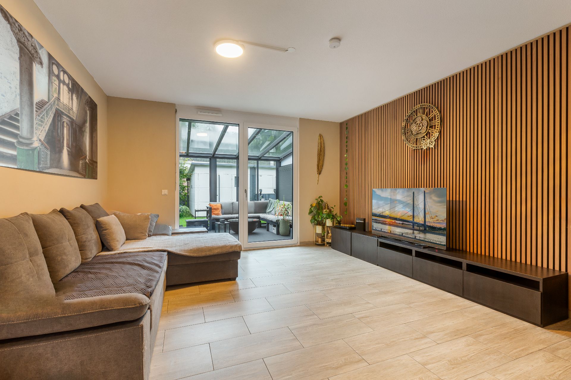 Modernes Wohnzimmer mit Ecksofa, Fernseher, Holzlamellenwand und Zugang zur Terrasse.
