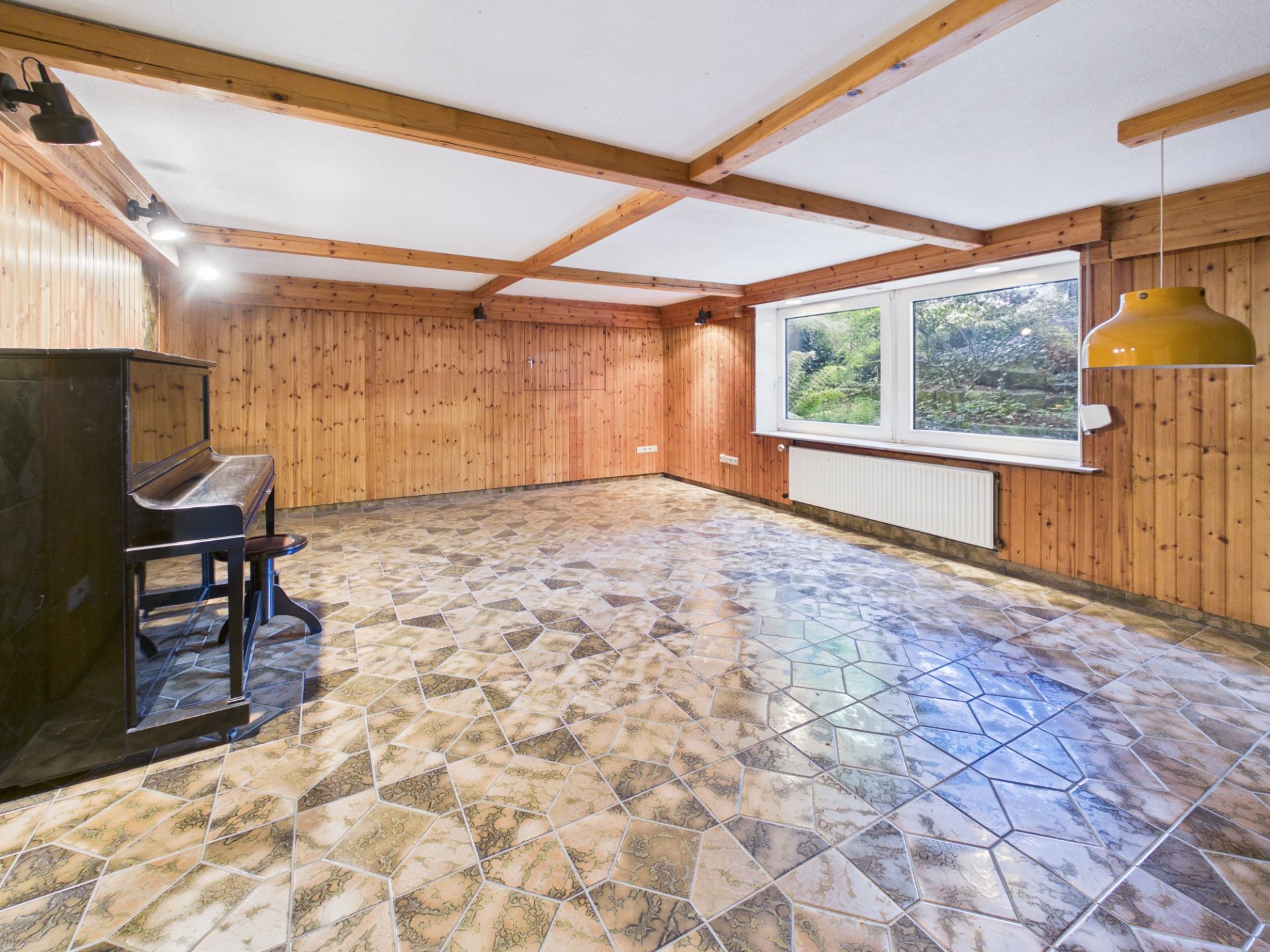 Großer Raum mit Fliesenboden, Holzvertäfelung, Fenster, Klavier und gelber Lampe.
