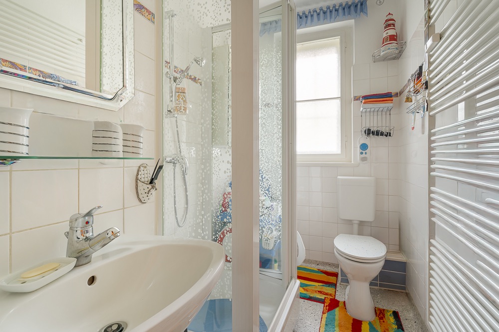 Helles Badezimmer mit Waschbecken, Dusche, WC und Fenster, bunte Teppiche auf dem Boden.