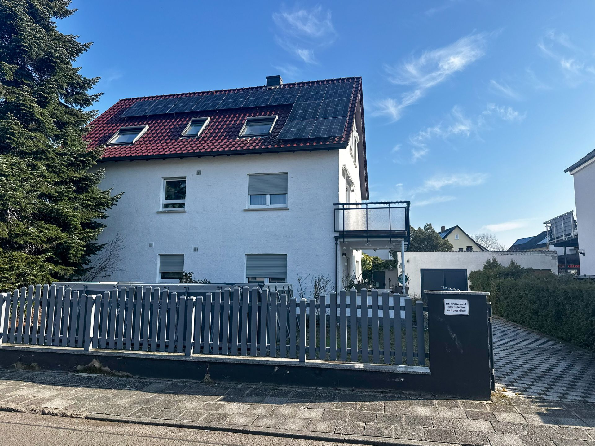 Weiße Wohnhaus mit rotem Dach und Solarpanelen, Vorgarten und grauem Zaun.