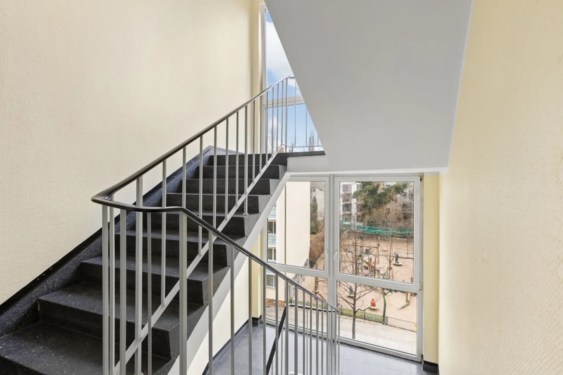Treppenhaus mit Geländer und großem Fenster mit Blick auf einen Spielplatz.