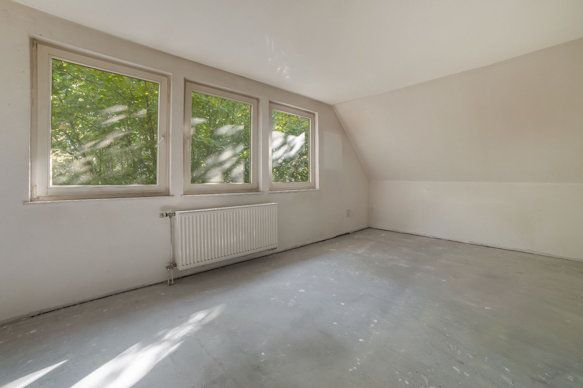 Leerer Raum mit drei Fenstern, weiß gestrichenen Wänden und einem Heizkörper unter den Fenstern.