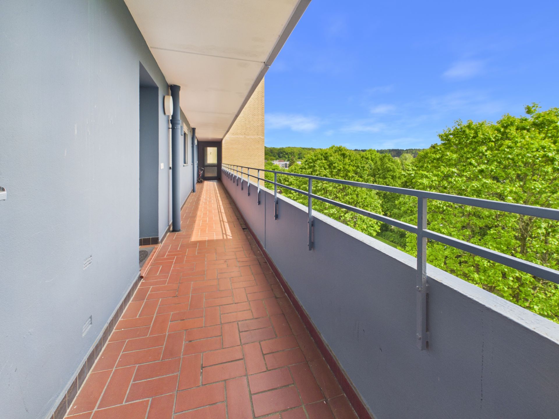 Langer Balkon mit roten Fliesen und Metallgeländer, Blick auf grüne Bäume.