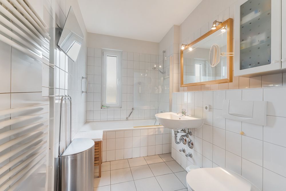 Helles Badezimmer mit Badewanne, Waschbecken, Spiegel und WC, weiße Fliesen an Wand und Boden.