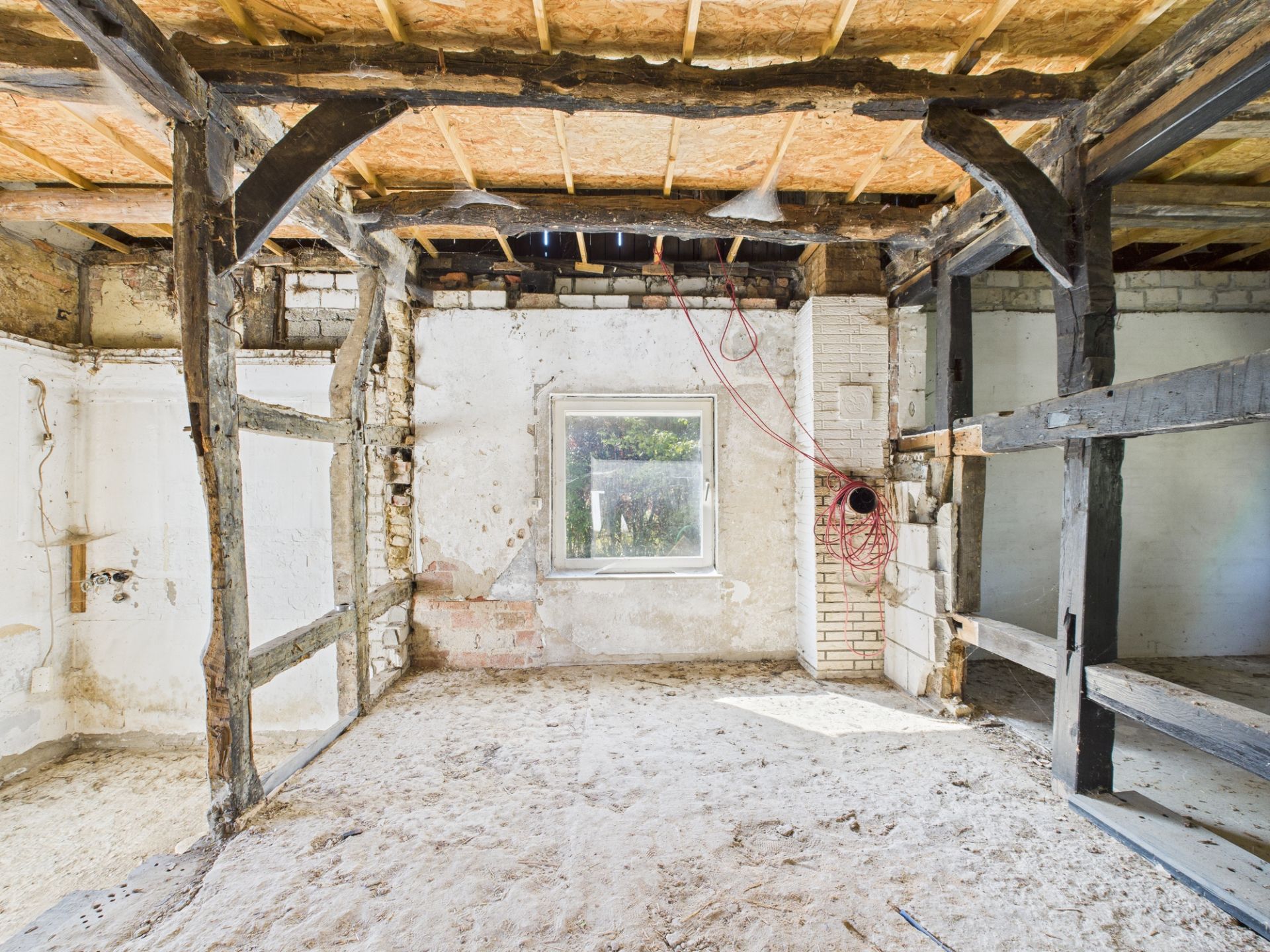 Innenraum eines Hauses im Rohbau mit freiliegenden Holzbalken und einem Fenster an der Rückwand.