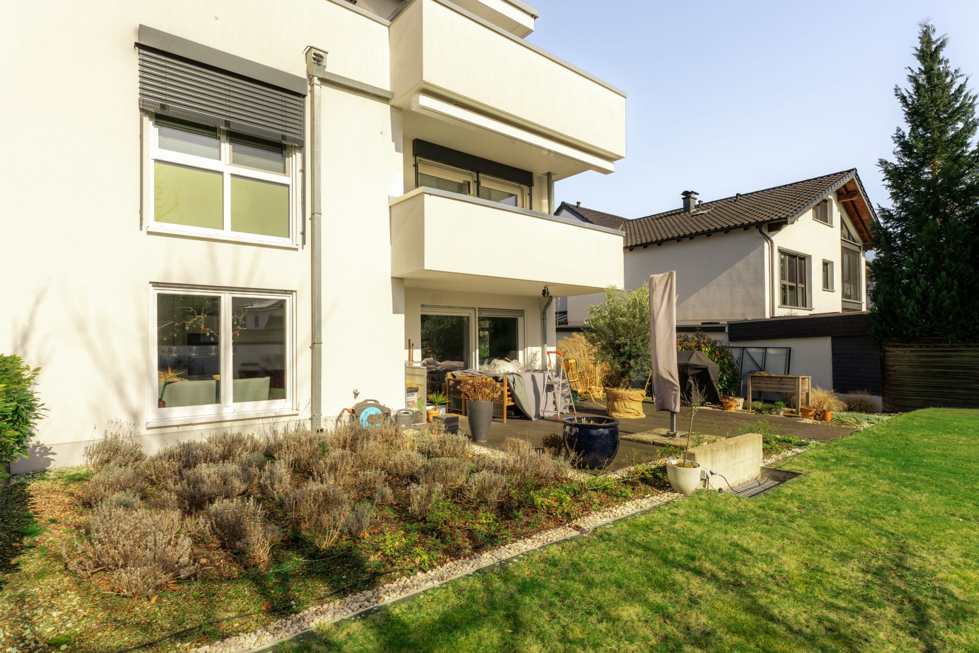 Moderne weiße Wohnhausfassade mit Balkon und Terrasse, angrenzend an einen Garten.
