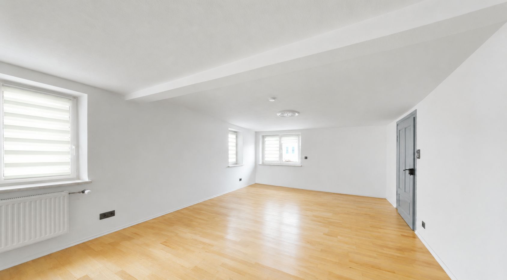 Heller Raum mit Parkettboden, weißen Wänden, drei Fenstern und grauer Tür.