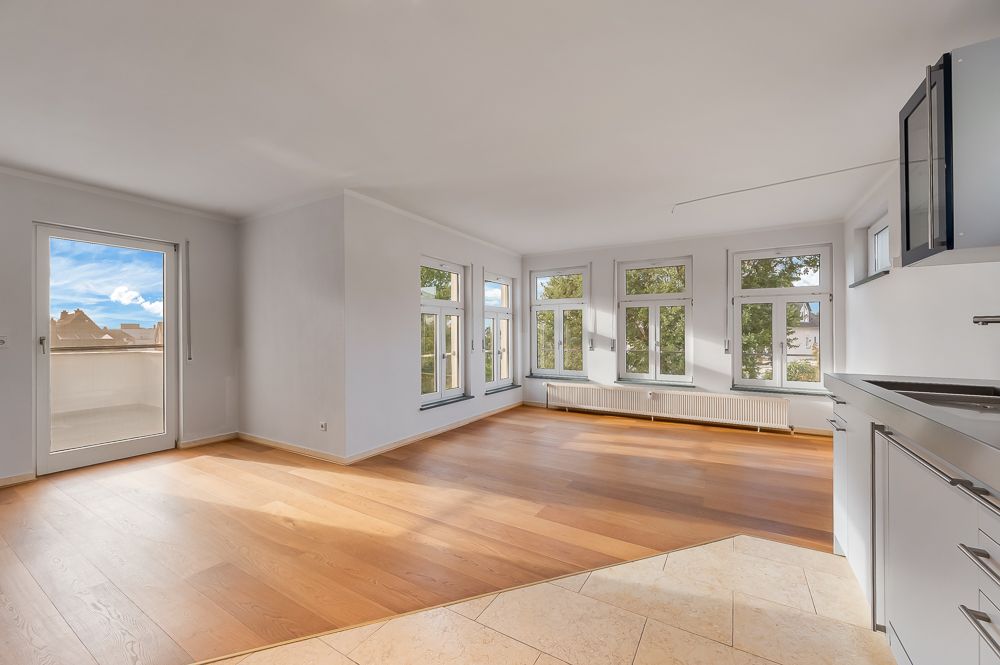 Helles Wohnzimmer mit Holzboden, vielen Fenstern und Zugang zu einem Balkon, offene Küchenzeile rechts.