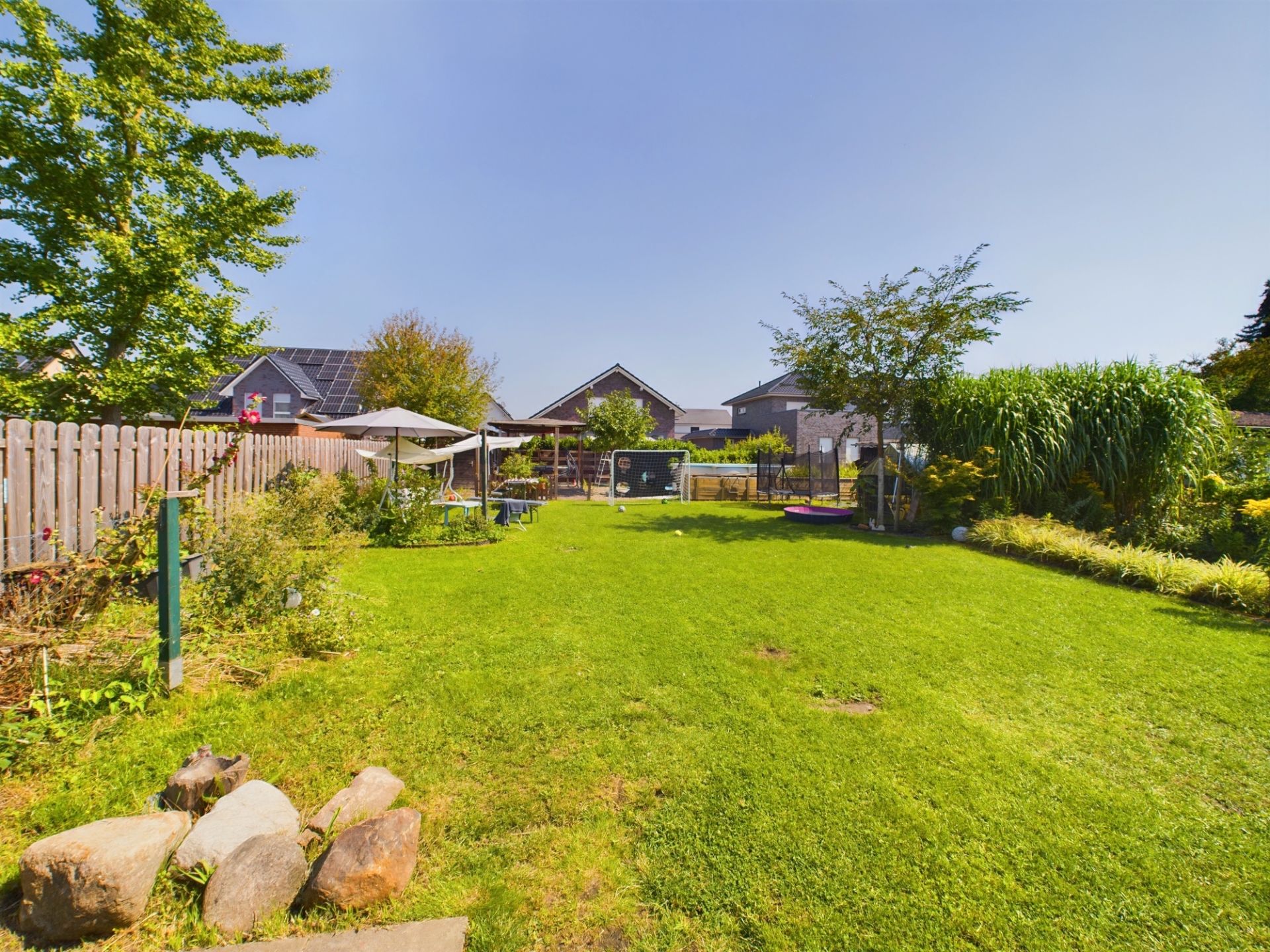 Großer grüner Garten mit Rasen, Bäumen, Zaun, Gartenmöbeln und einem Sonnenschirm.