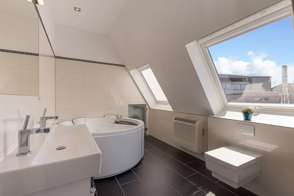 Modernes Badezimmer mit Eckbadewanne, Waschbecken, Wandtoilette und zwei Dachfenstern.