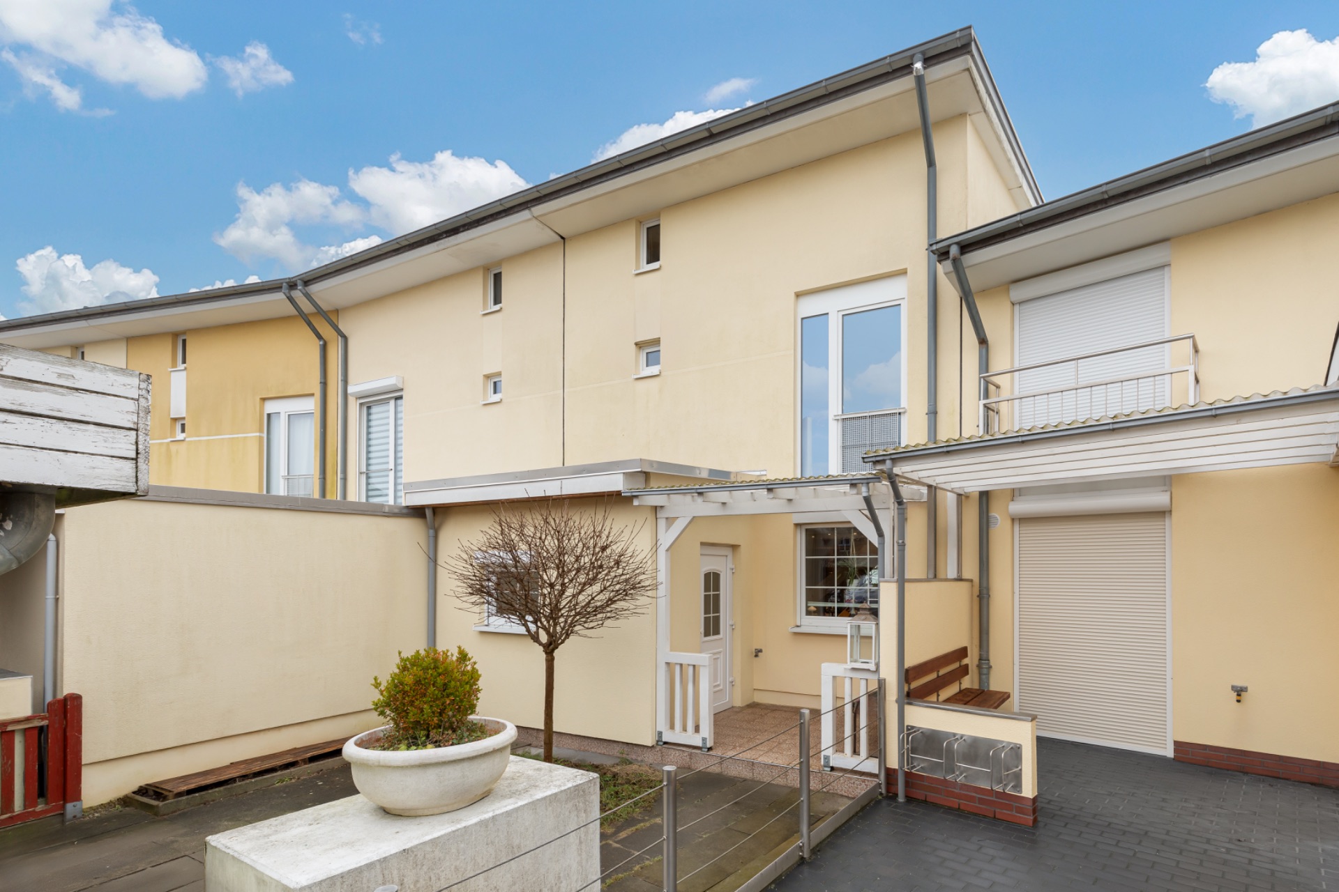 Modernes, gelbes Reihenhaus mit kleinem Garten, Terrasse, Baum im Topf und überdachtem Eingangsbereich.