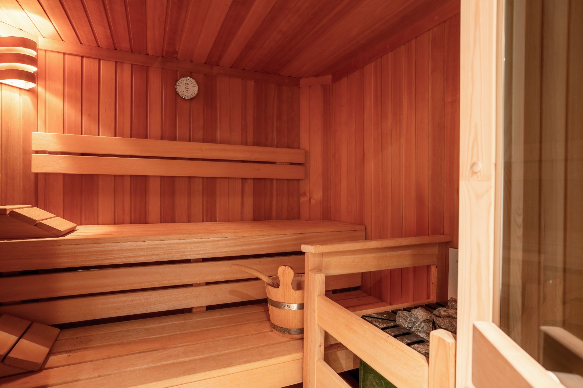 Innenraum einer Holzsauna mit Bank, Eimer, Steinen und Wanduhr.