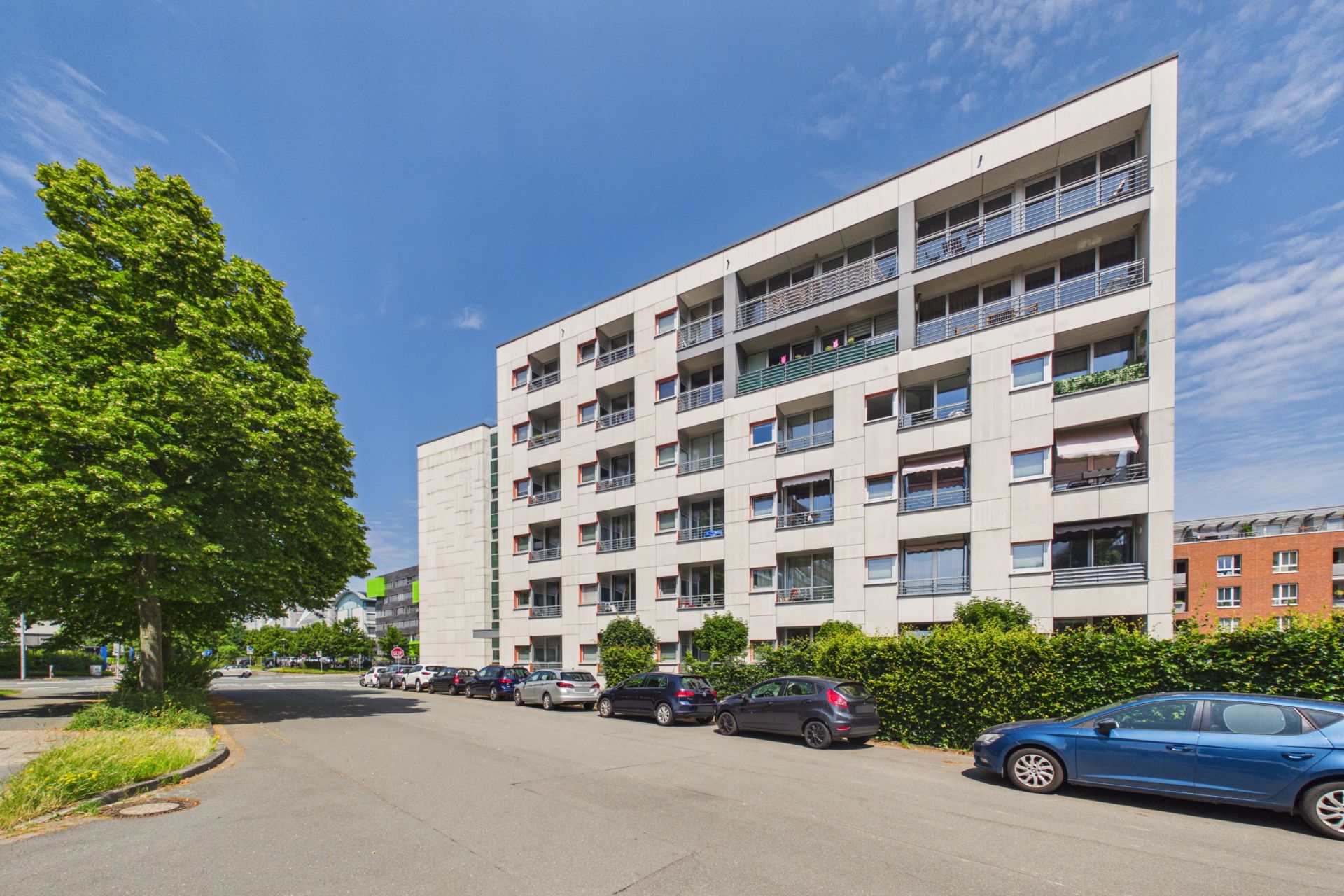 Mehrstöckiges Wohngebäude mit Balkonen und geparkten Autos an der Straße bei sonnigem Wetter.