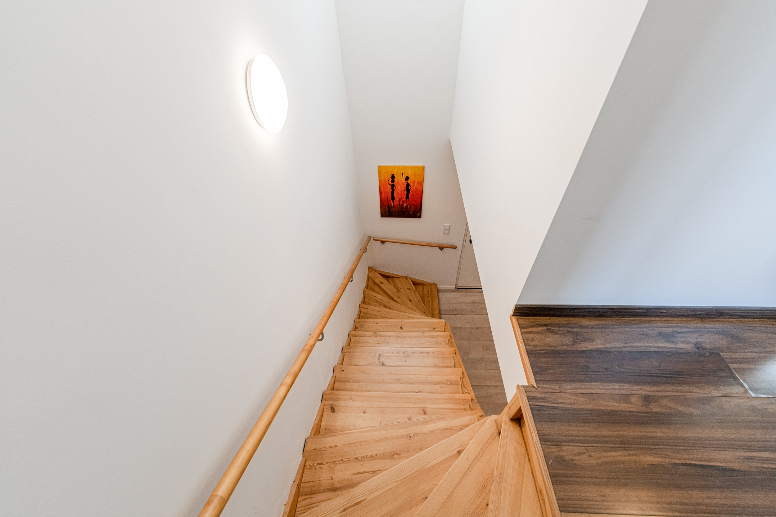 Holztreppe mit Handlauf, weiß gestrichene Wände, Bild an der Wand und Deckenleuchte.