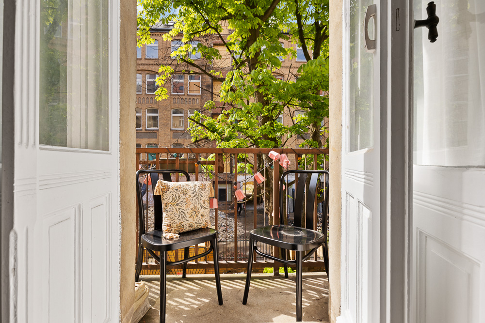 Kleiner Balkon mit zwei Stühlen, einem Tisch und Blick auf Bäume sowie ein Wohngebäude.
