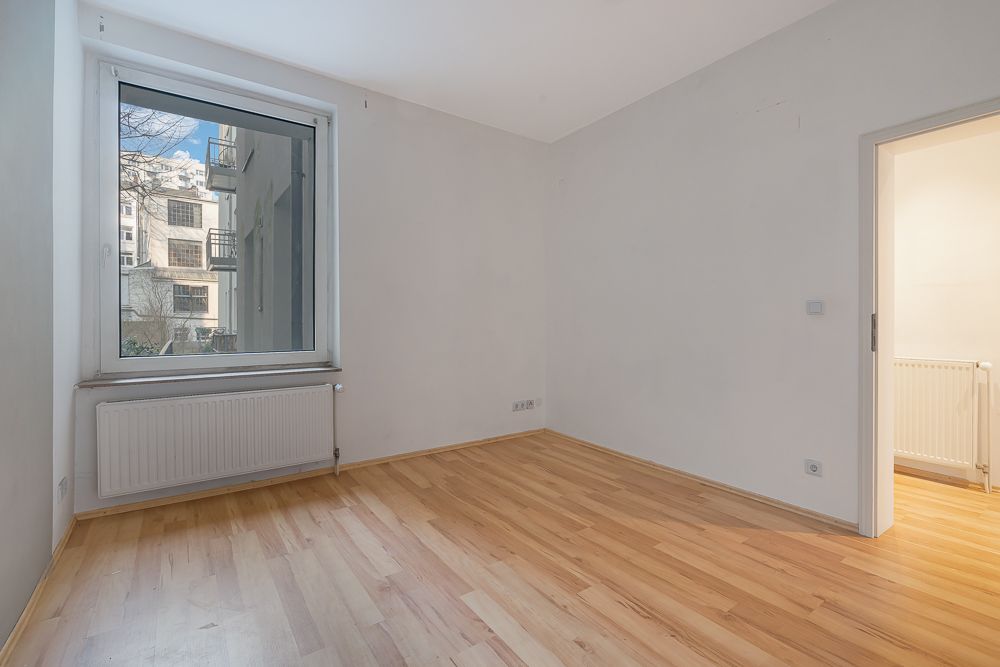 Leeres Zimmer mit Holzboden, großem Fenster und Heizung unter dem Fenster.
