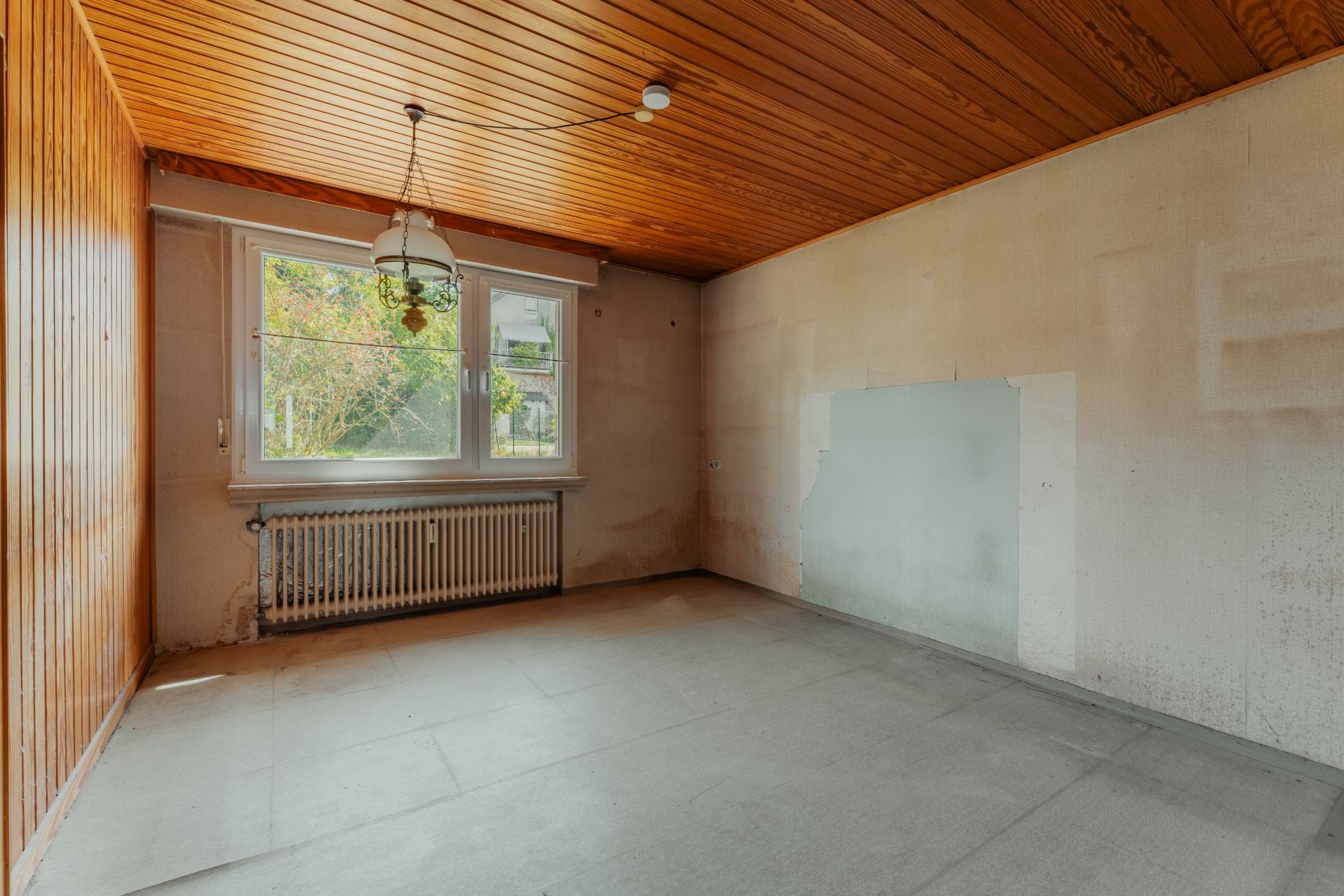 Leerer Raum mit Holzdecke, großem Fenster und Heizkörper unter dem Fenster.