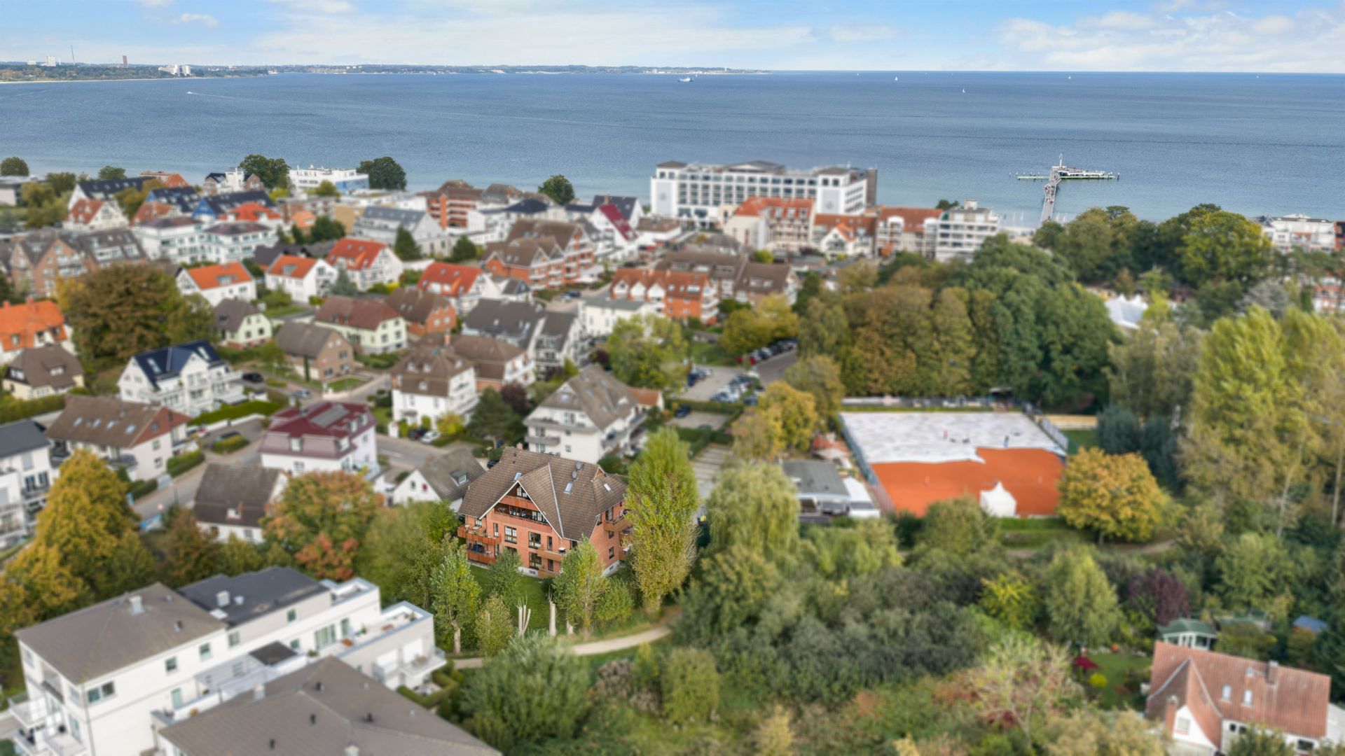 Luftaufnahme eines Wohngebiets mit Häusern, Bäumen und Blick auf das Meer im Hintergrund.