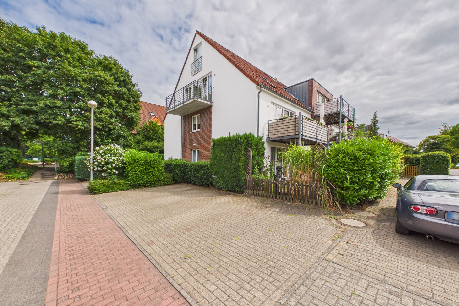 Modernes Wohnhaus mit Balkonen, Hecken und angrenzendem Parkplatz bei bewölktem Himmel.