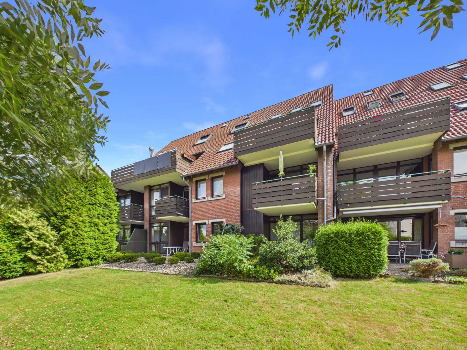 Mehrfamilienhaus mit Balkonen, rotem Dach und begrüntem Garten im Vordergrund.