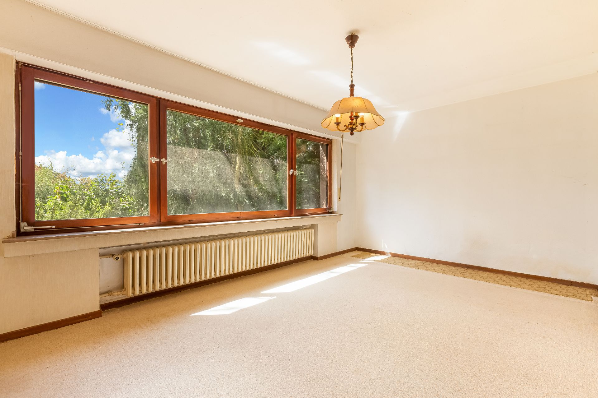 Helles, leeres Zimmer mit großem Fenster, Heizkörper und Deckenlampe.