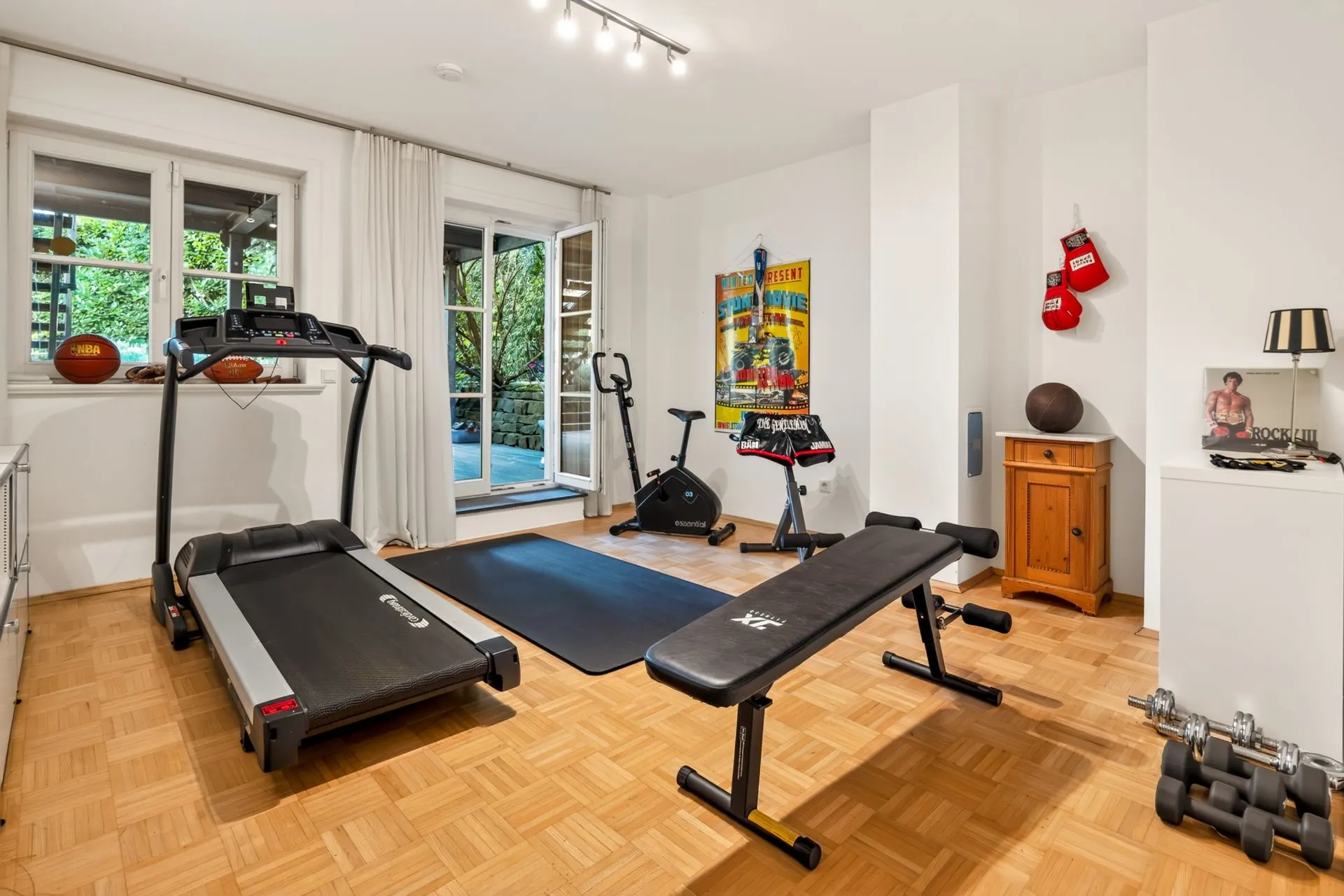 Helles Fitnesszimmer mit Laufband, Hantelbank, Fahrradtrainer und Hanteln auf Parkettboden.
