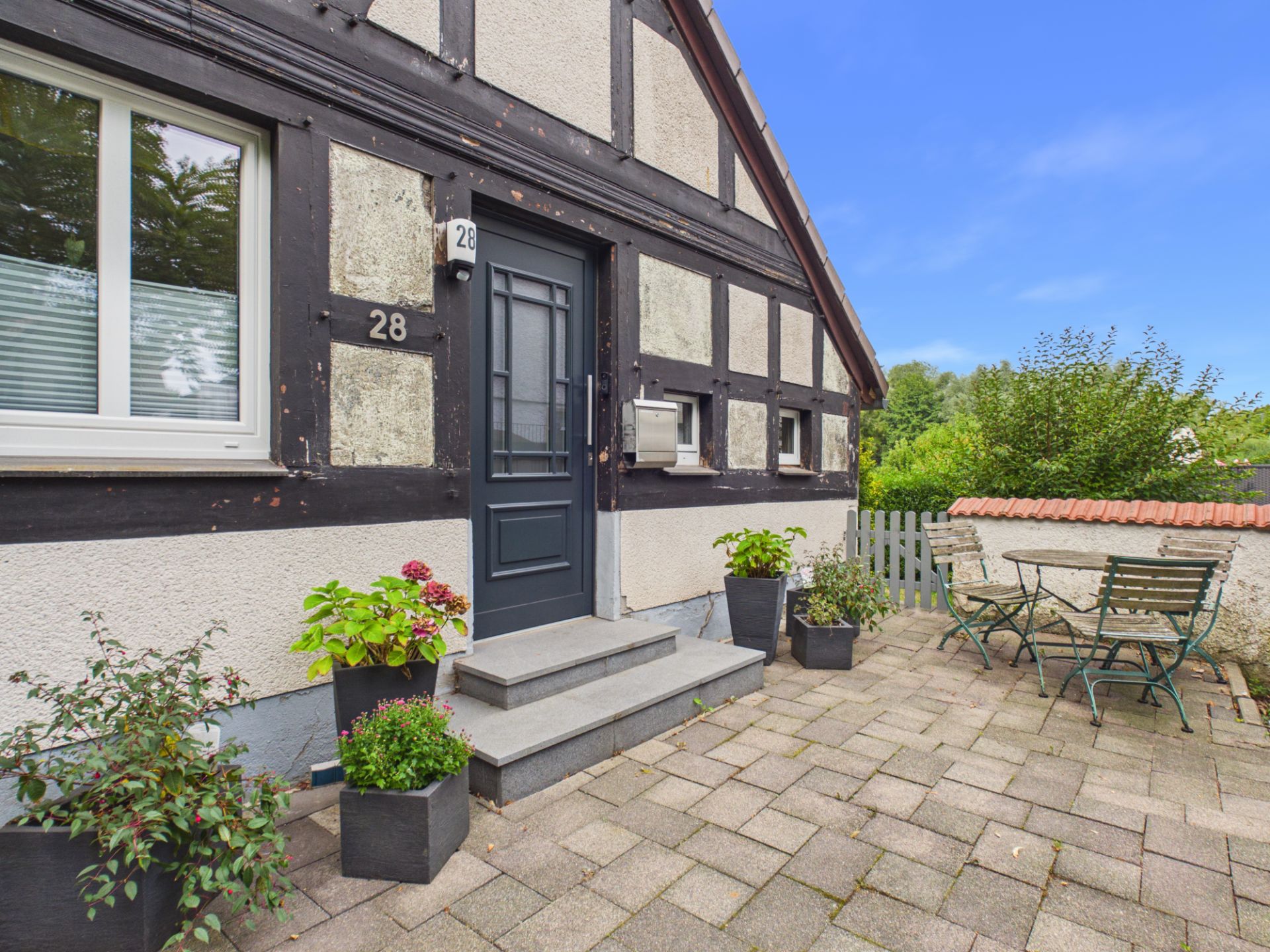 Eingangsbereich eines Fachwerkhauses mit Pflanzen und Sitzgruppe auf gepflasterter Terrasse.