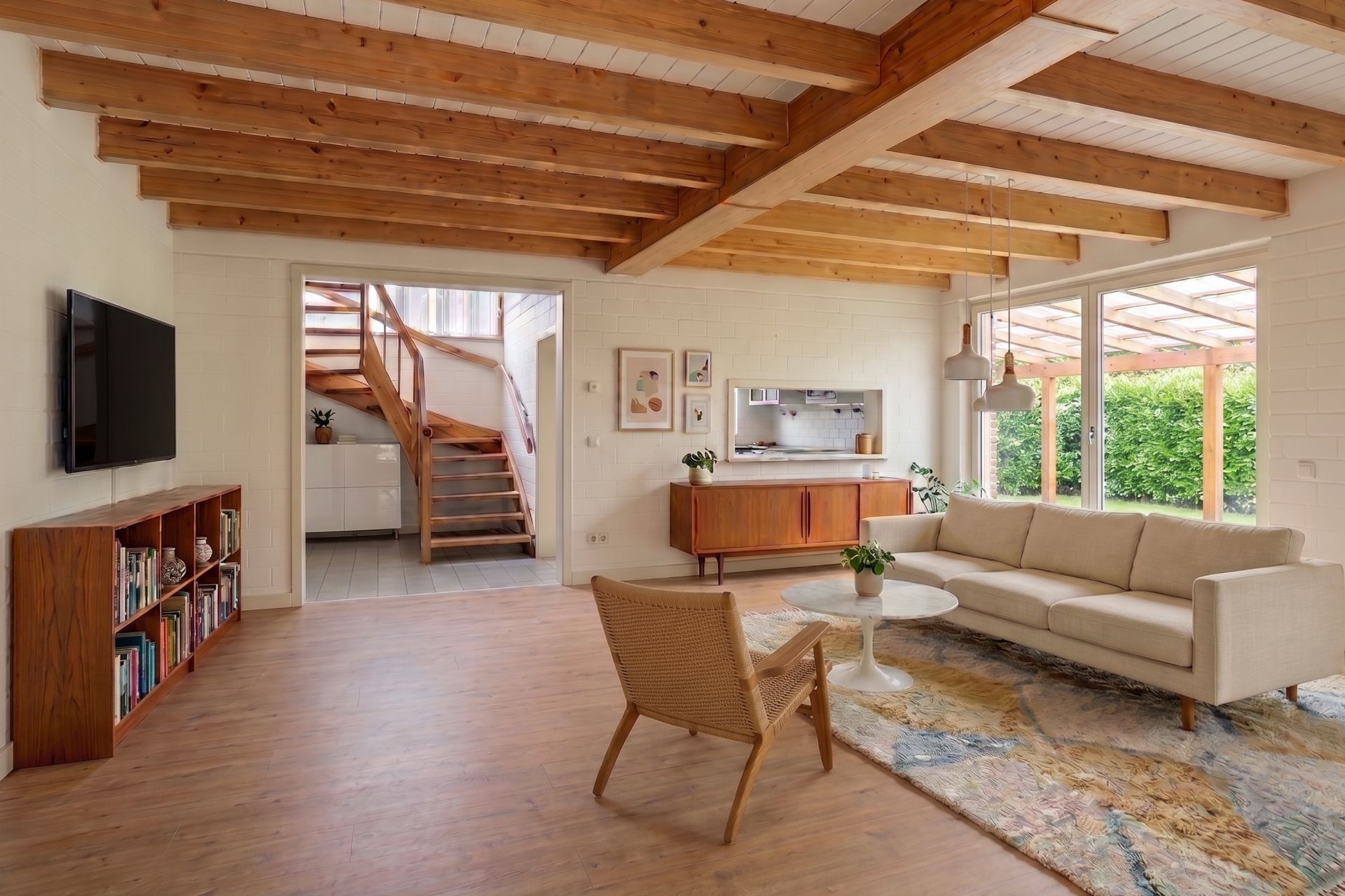 Helles Wohnzimmer mit Holzbalkendecke, Sofa, Sessel, Regal und Blick auf eine Holztreppe.