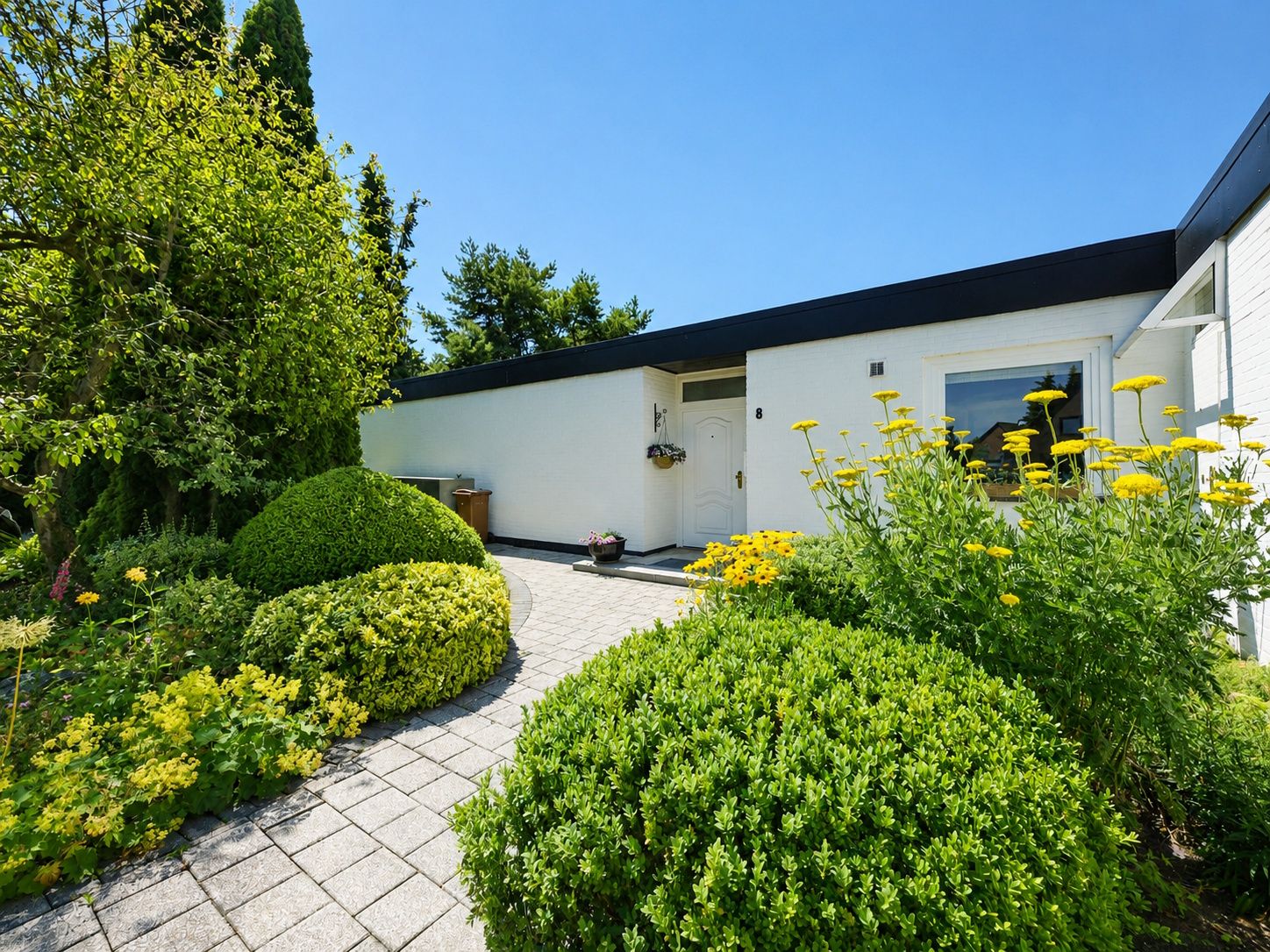 Eingang eines weißen Hauses mit gepflegtem Vorgarten und blühenden gelben Blumen.
