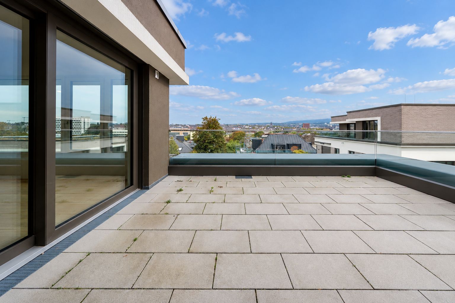 Große Dachterrasse mit Fliesen, Glasgeländer und Blick auf umliegende Gebäude bei Tageslicht.