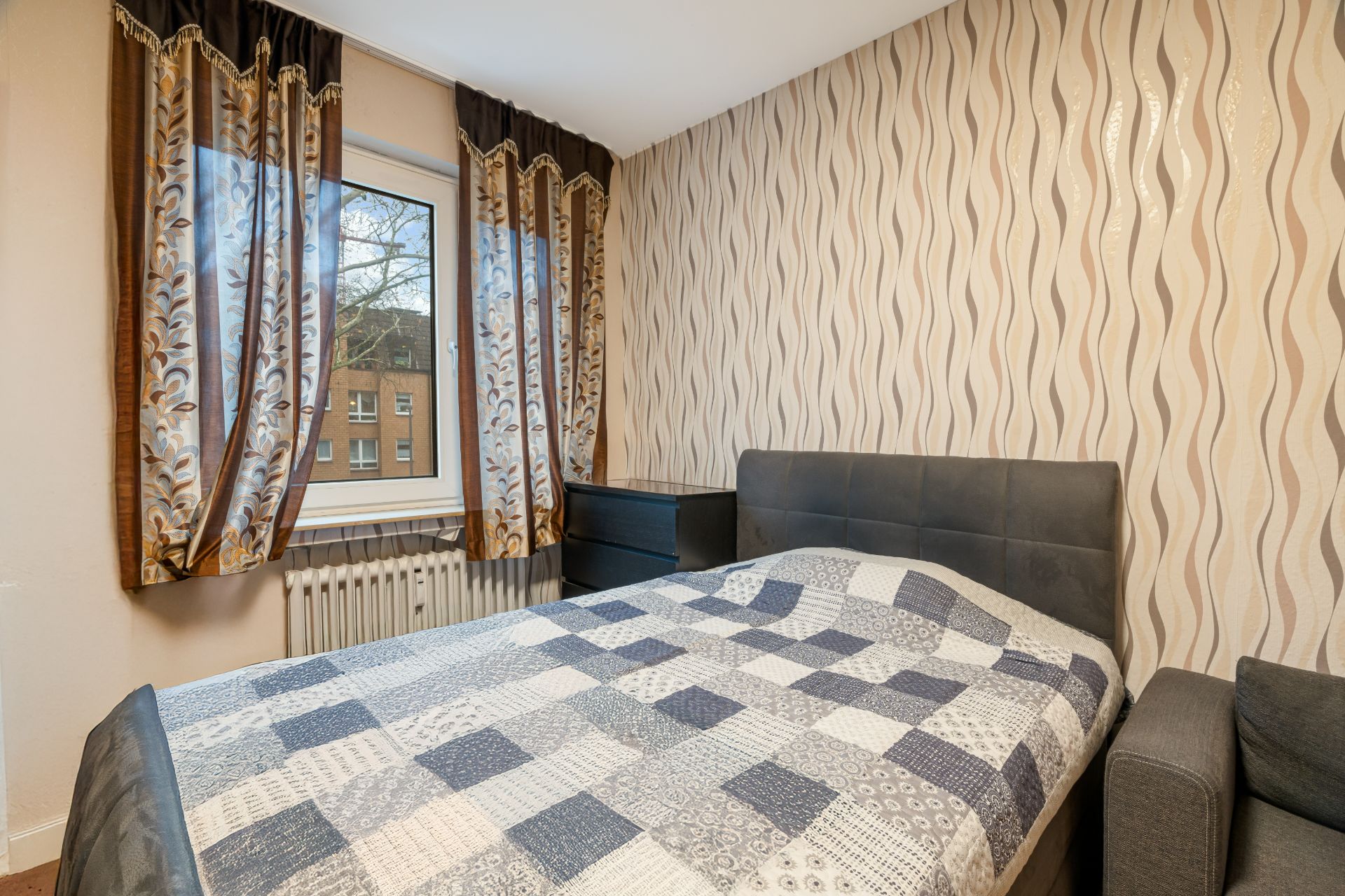 Schlafzimmer mit Doppelbett, gemusterten Vorhängen und wellenförmiger Tapete.