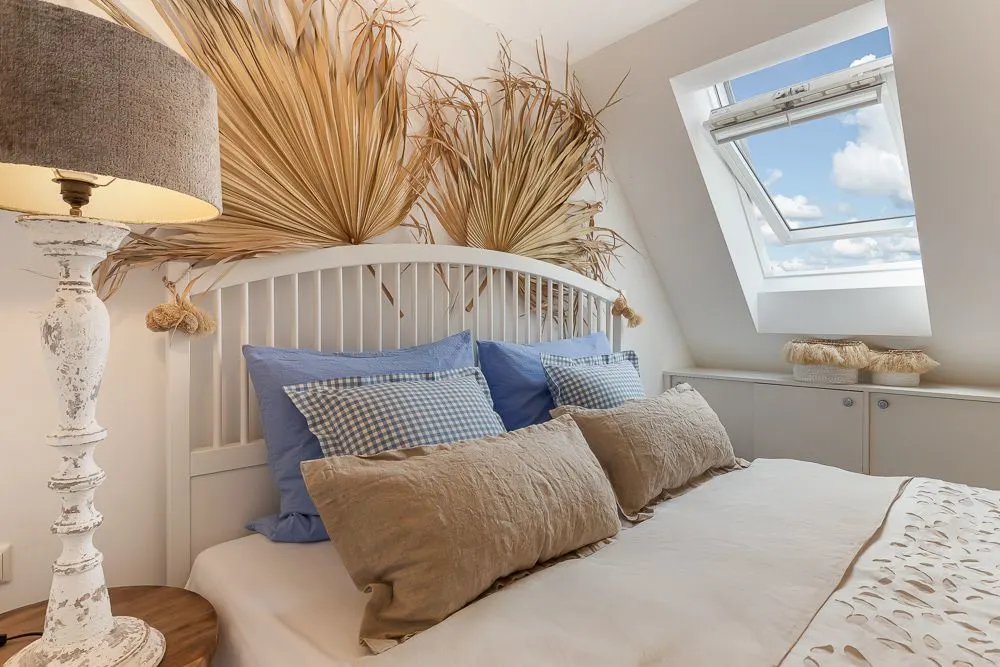 Helles Schlafzimmer mit Bett, blauen Kissen, Dekozweigen und offenem Dachfenster.