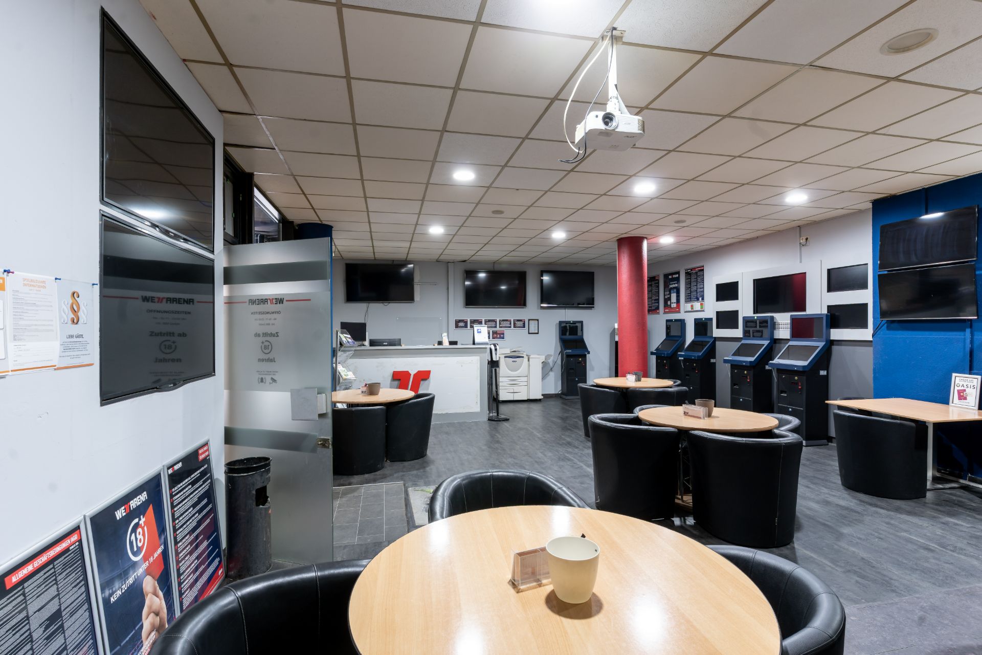 Runder Holztisch mit schwarzen Stühlen in einem Raum mit mehreren Fernsehern und Automaten an den Wänden.