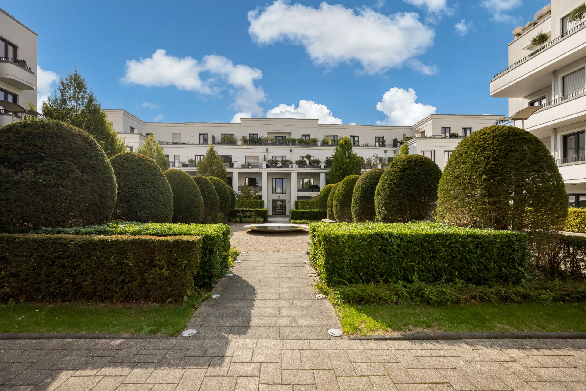 Gepflegter Innenhof mit Hecken, runden Büschen und einem Springbrunnen, umgeben von Wohngebäuden.