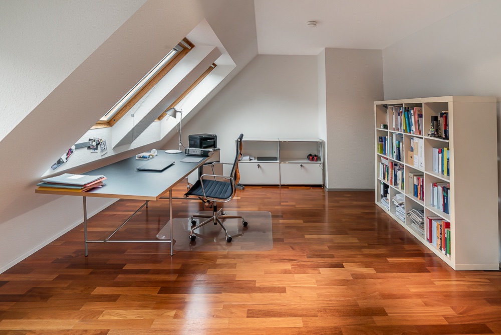 Helles Arbeitszimmer mit Schreibtisch, Stuhl, Bücherregal und Dachfenstern auf Holzfußboden.