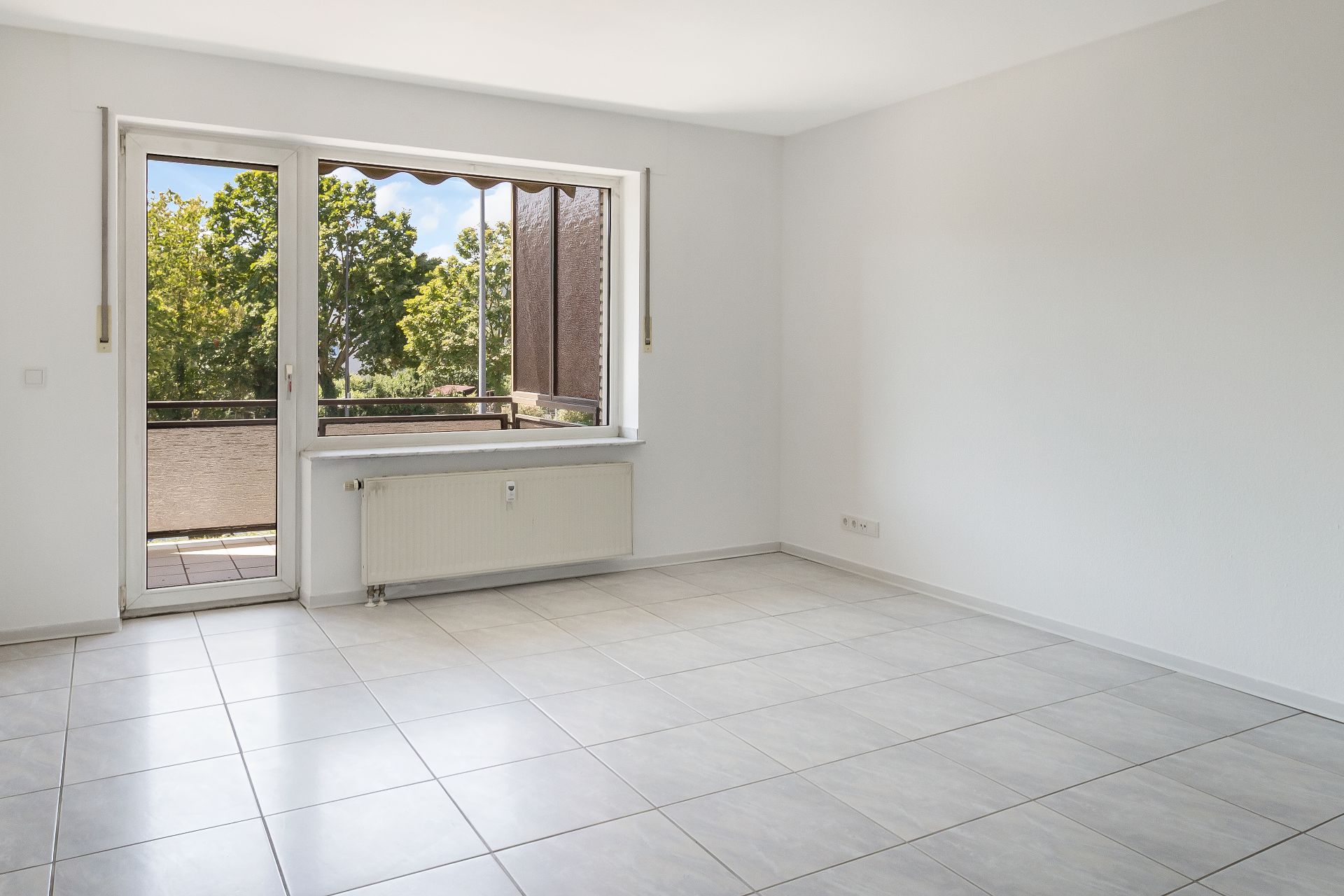 Helles, leeres Zimmer mit Fliesenboden und Zugang zu einem Balkon mit Blick auf Bäume.
