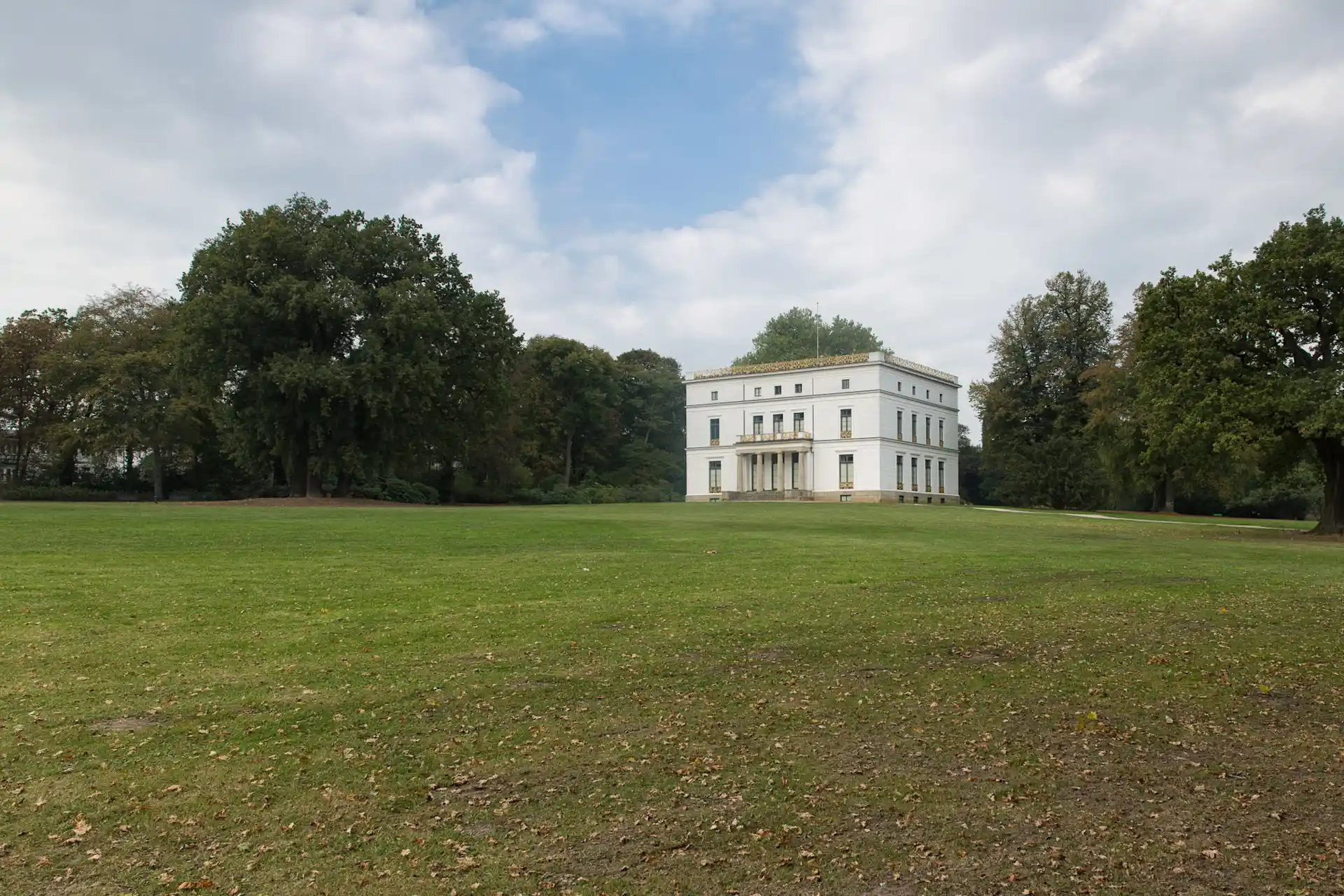 Großes weißes Haus mit Säulen, umgeben von Rasen und Bäumen unter bewölktem Himmel.