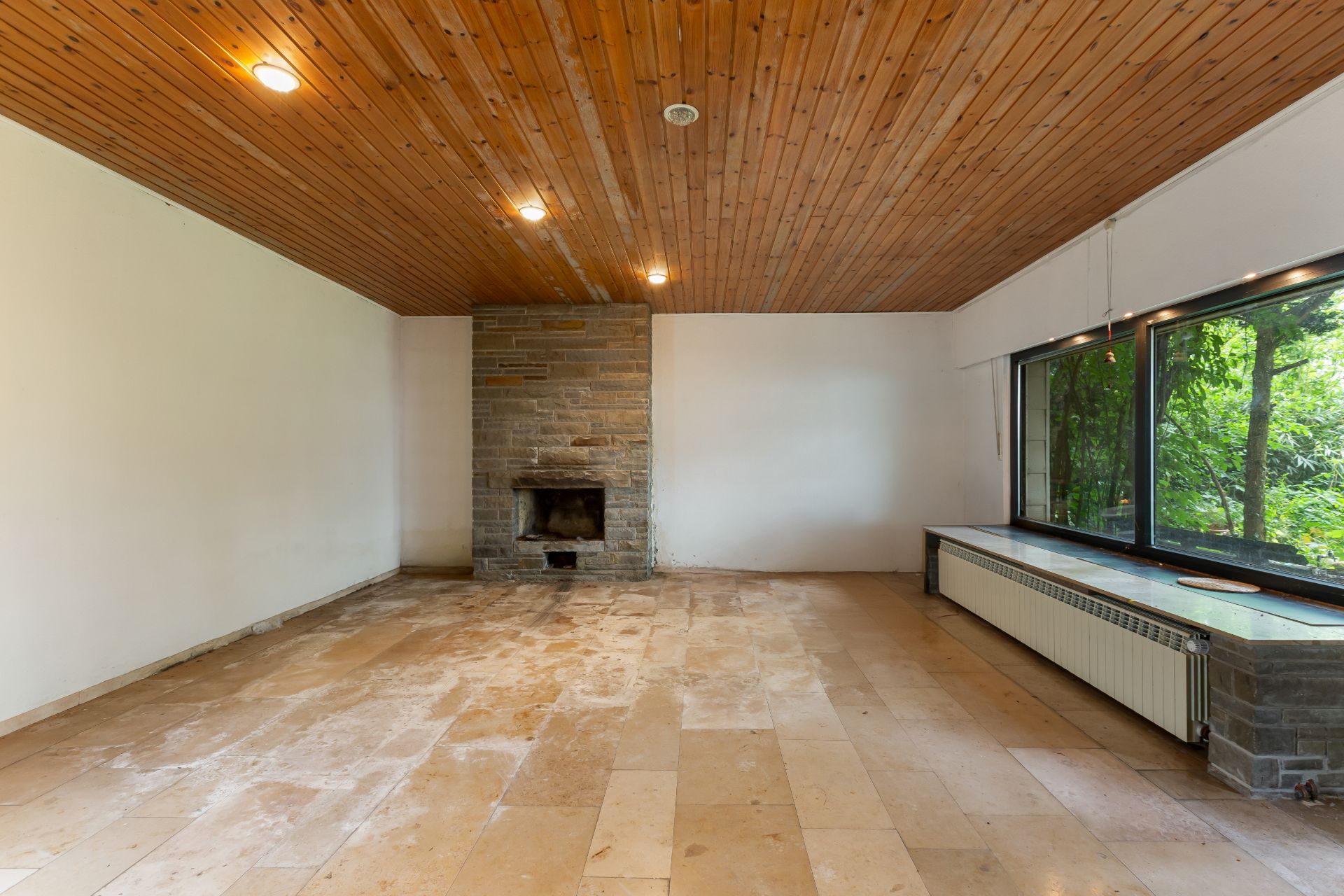 Leeres Wohnzimmer mit Holzbalkendecke, Kamin und großem Fenster mit Blick ins Grüne.