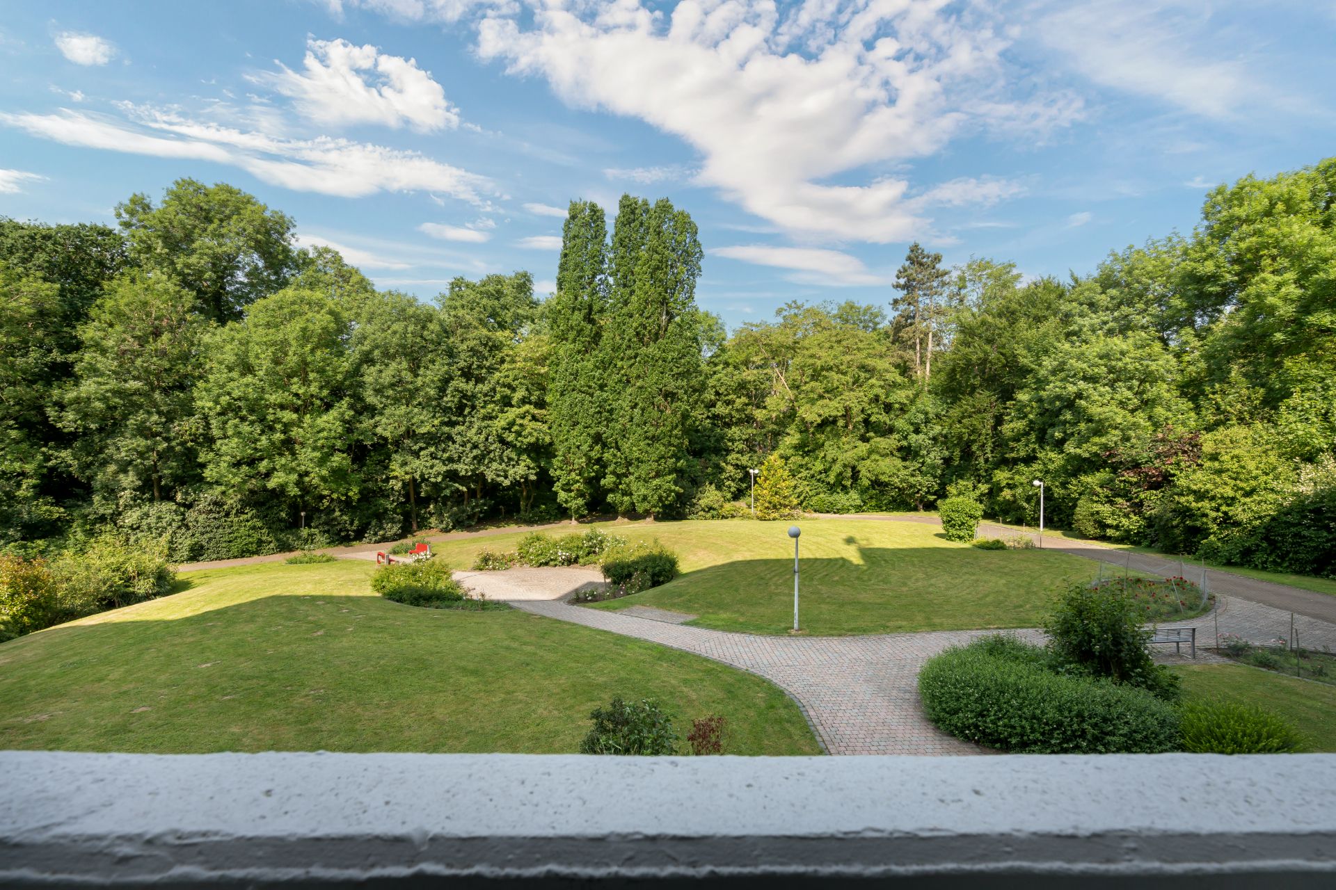 Blick vom Balkon auf eine gepflegte grüne Gartenanlage mit Wegen und vielen Bäumen.