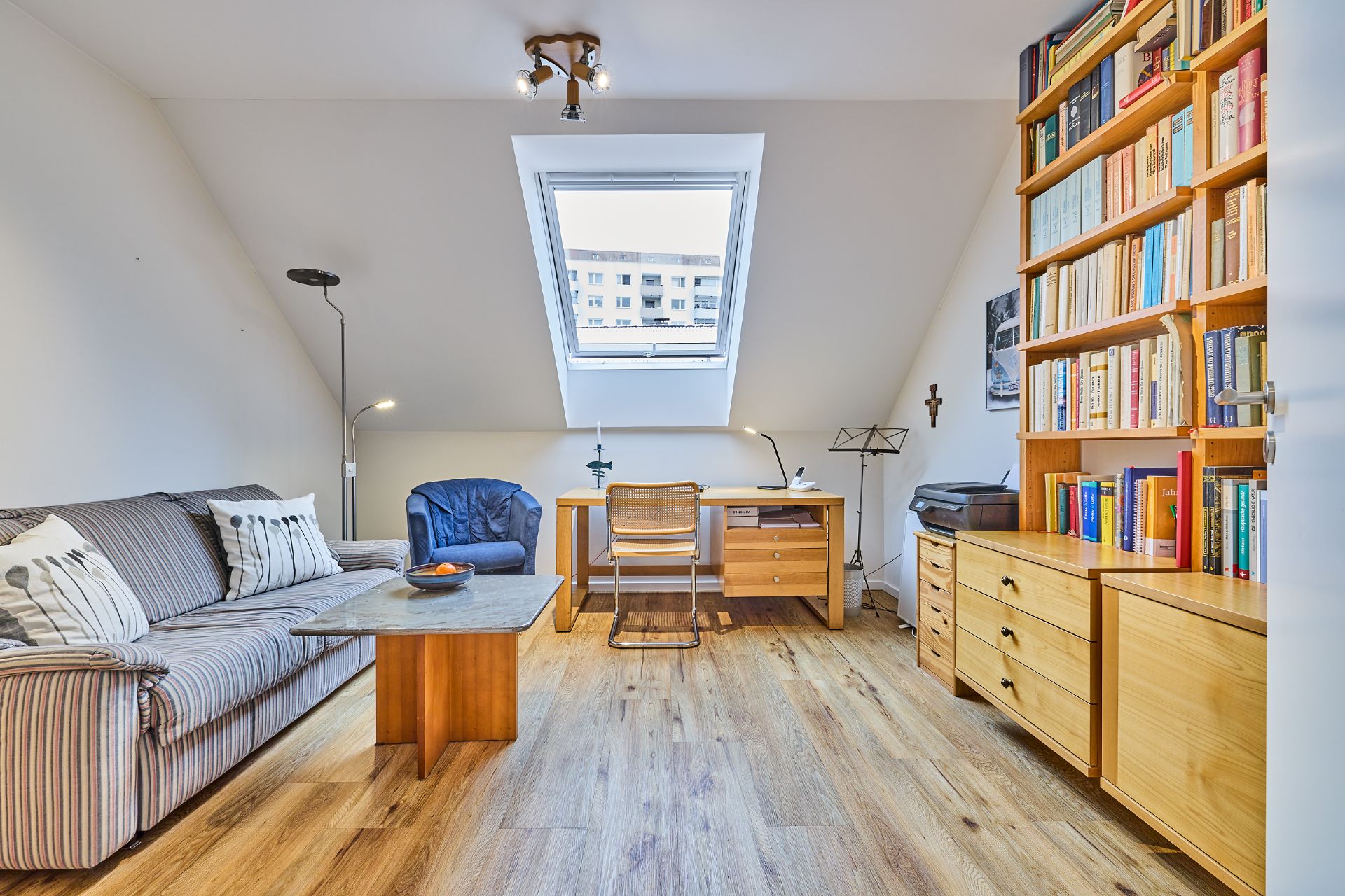 Wohnzimmer mit Sofa, Schreibtisch, Bücherregal und großem Fenster unter dem Dach.