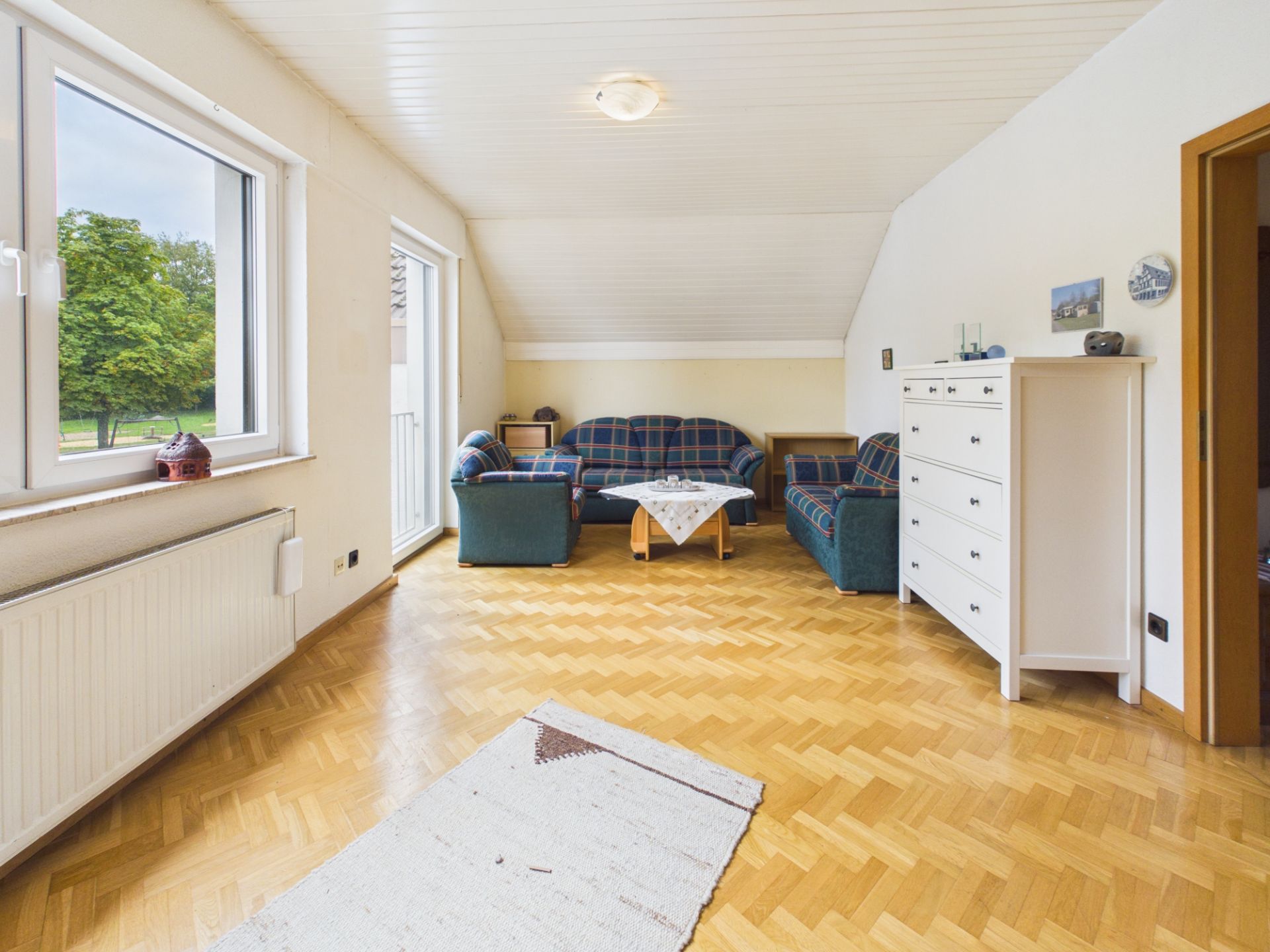 Helles Wohnzimmer mit Parkettboden, Sofas, Kommode und großem Fenster mit Blick ins Grüne.