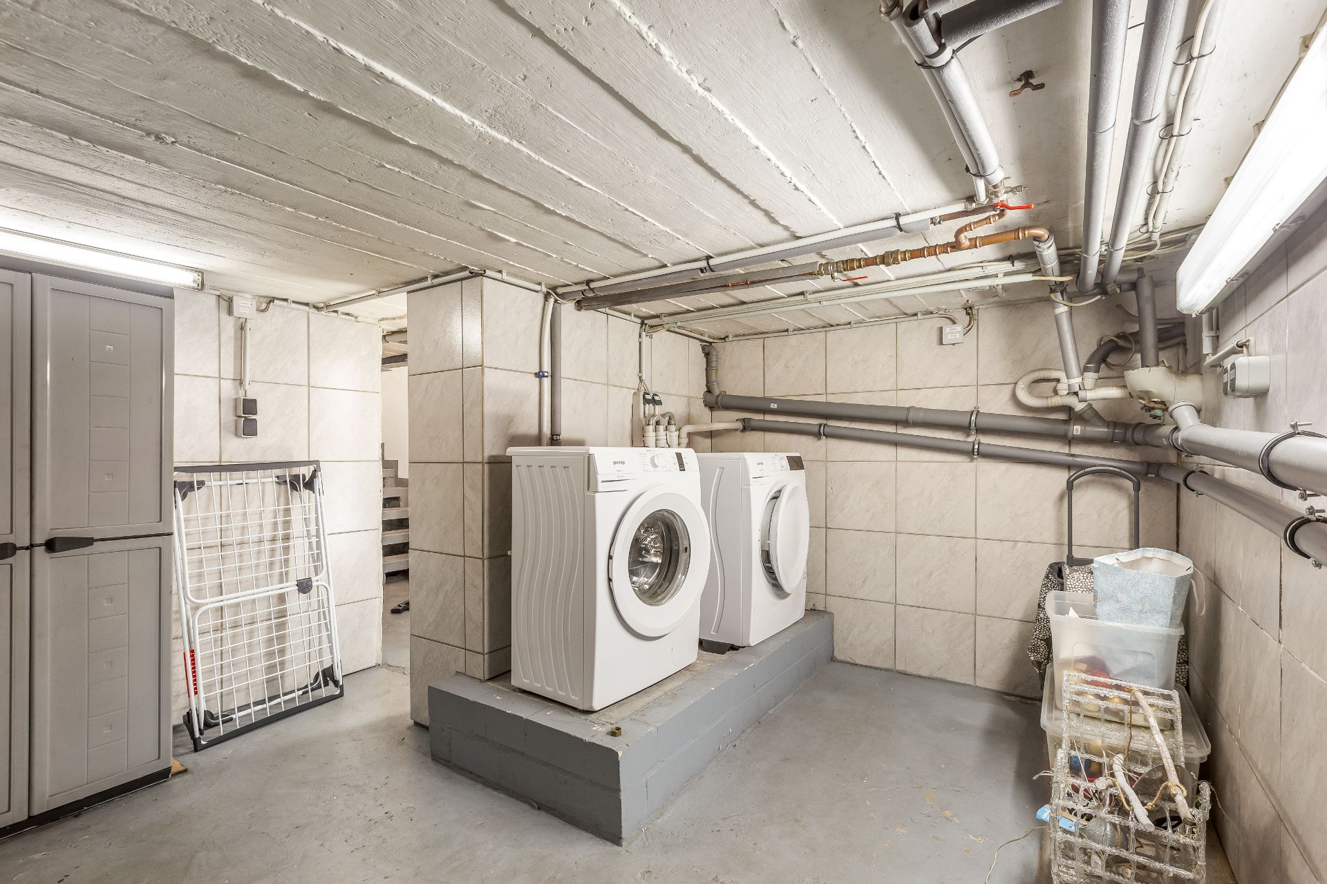 Waschraum mit zwei Waschmaschinen, Rohren an der Decke und einem Aufbewahrungsschrank.