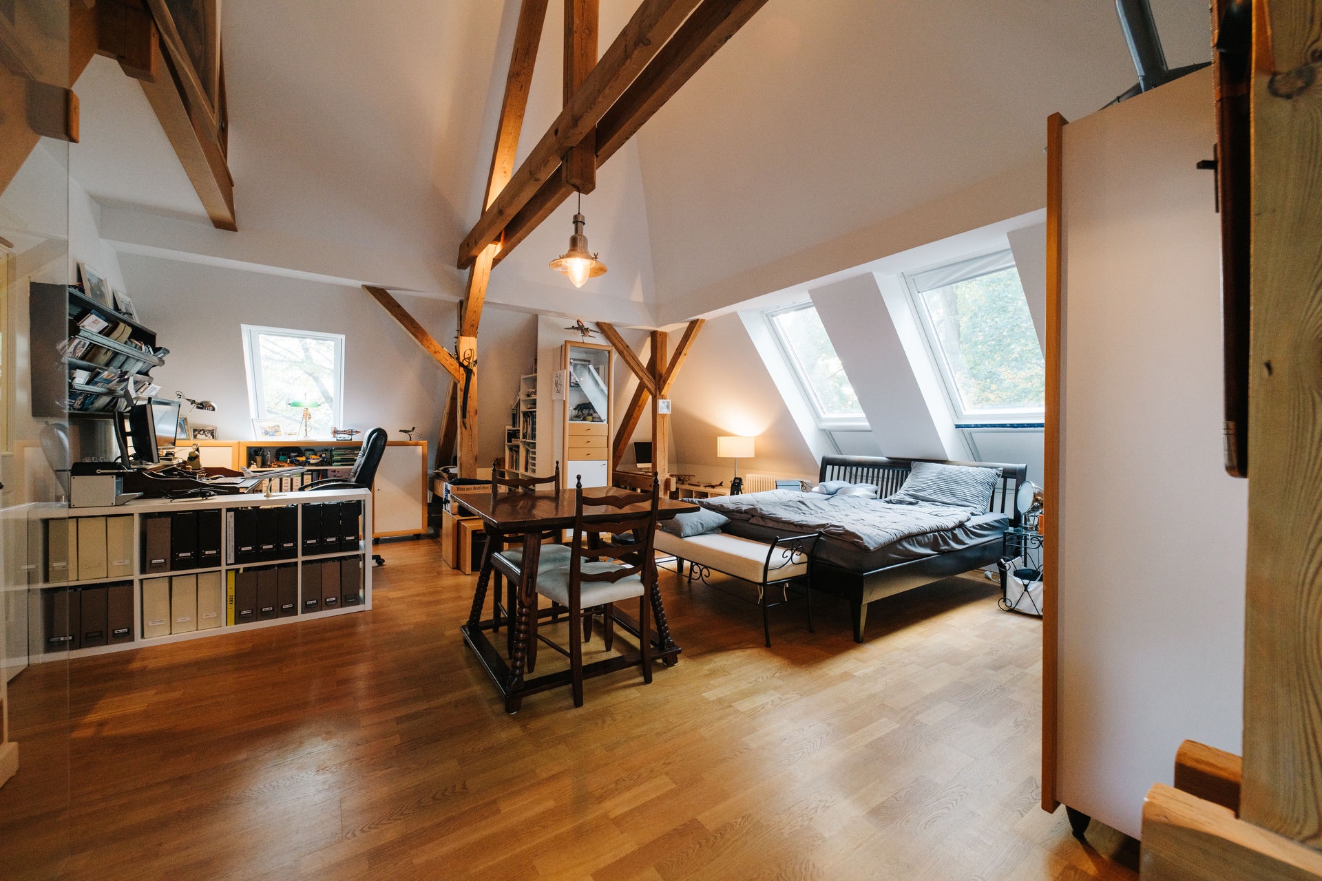 Großer Wohnraum mit Holzboden, Schreibtisch, Esstisch, Bett und sichtbaren Deckenbalken.