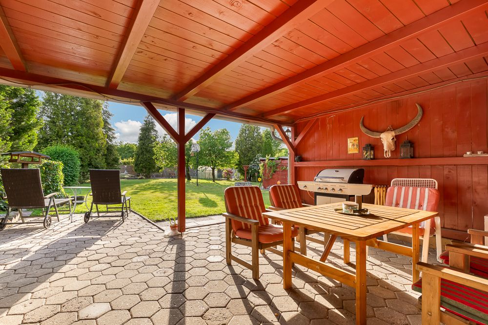Überdachte Terrasse mit Holztisch, Grill und Gartenblick, daneben zwei Liegestühle auf Steinboden.