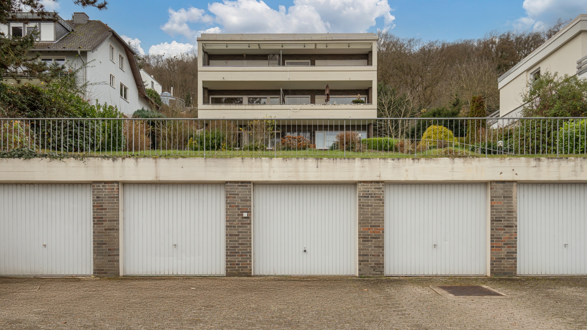 Mehrfamilienhaus mit Balkonen hinter vier geschlossenen Garagentoren und begrünter Böschung.