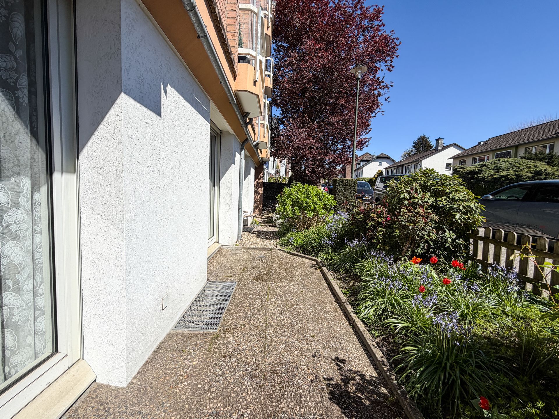 Gepflasterter Weg neben einem Haus mit Blumenbeeten und Bäumen im Vorgarten.
