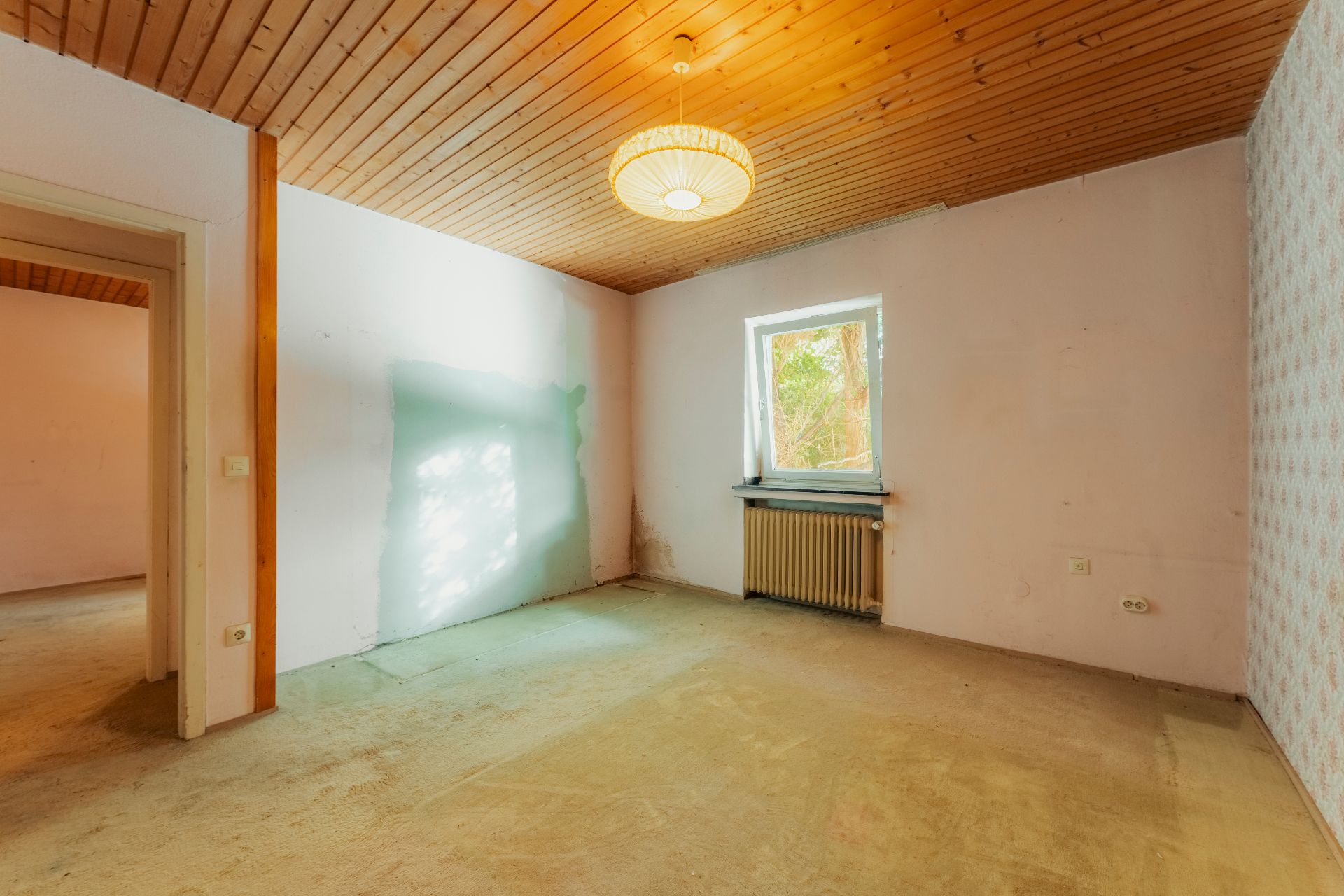 Leerer Raum mit Holzbalkendecke, Fenster, Heizung und Lampe, keine Möbel.