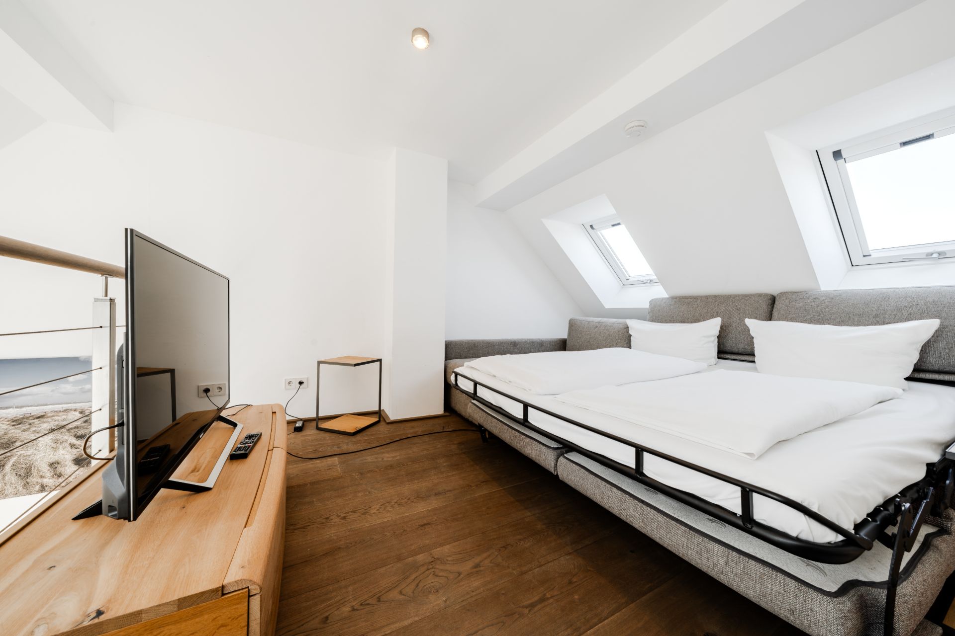 Helles Schlafzimmer mit grauem Sofa-Bett, Fernseher auf Holztisch und zwei Dachfenstern.