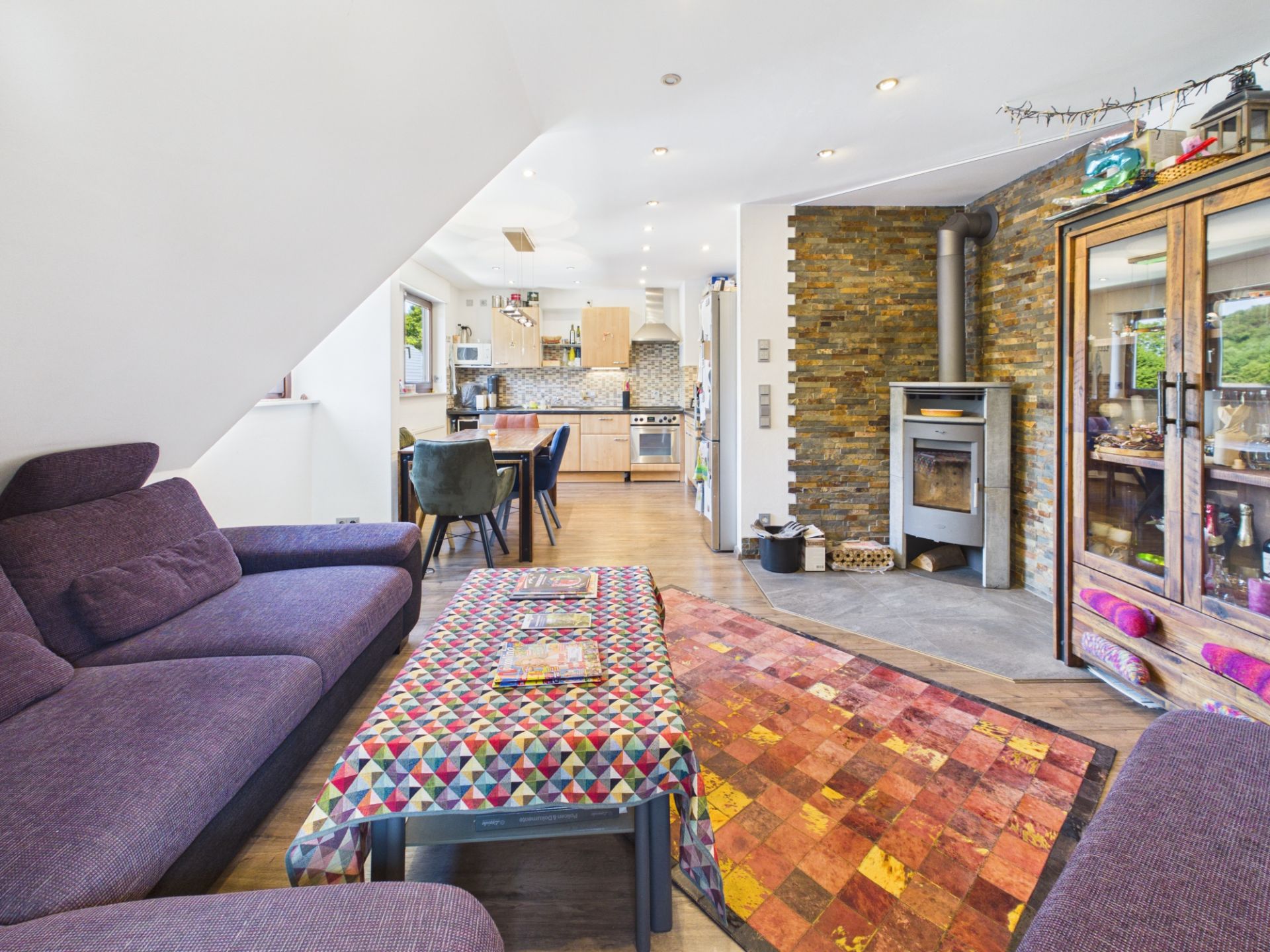 Helles Wohnzimmer mit lila Sofa, buntem Teppich, Esstisch, Kaminofen und offener Küche.